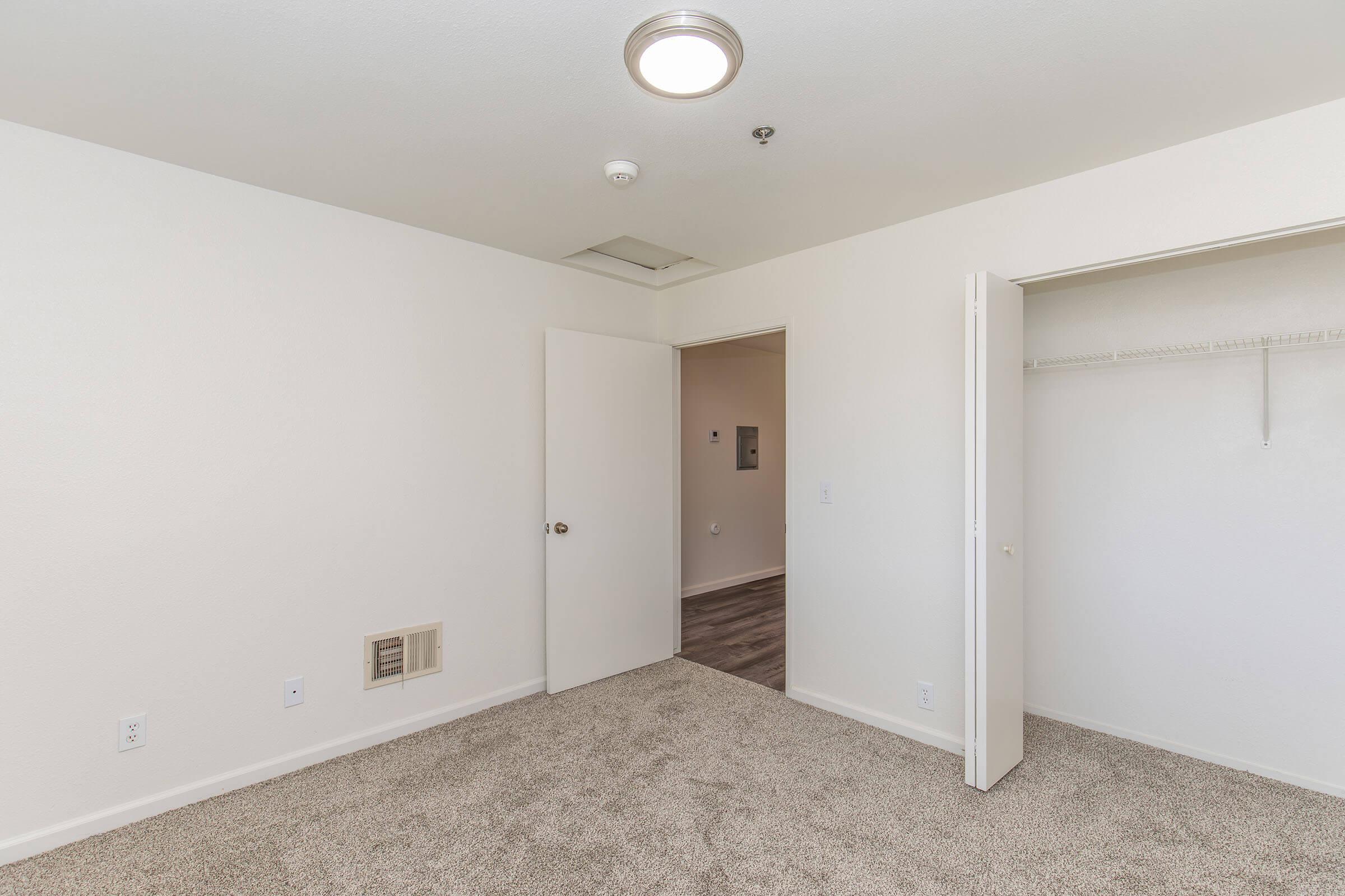A small, empty room with light beige walls and carpet flooring. There is a ceiling light fixture, a closed door leading to another space, and an open closet with a hanging rod. The room appears well-lit and clean, suitable for various uses such as a bedroom or office.
