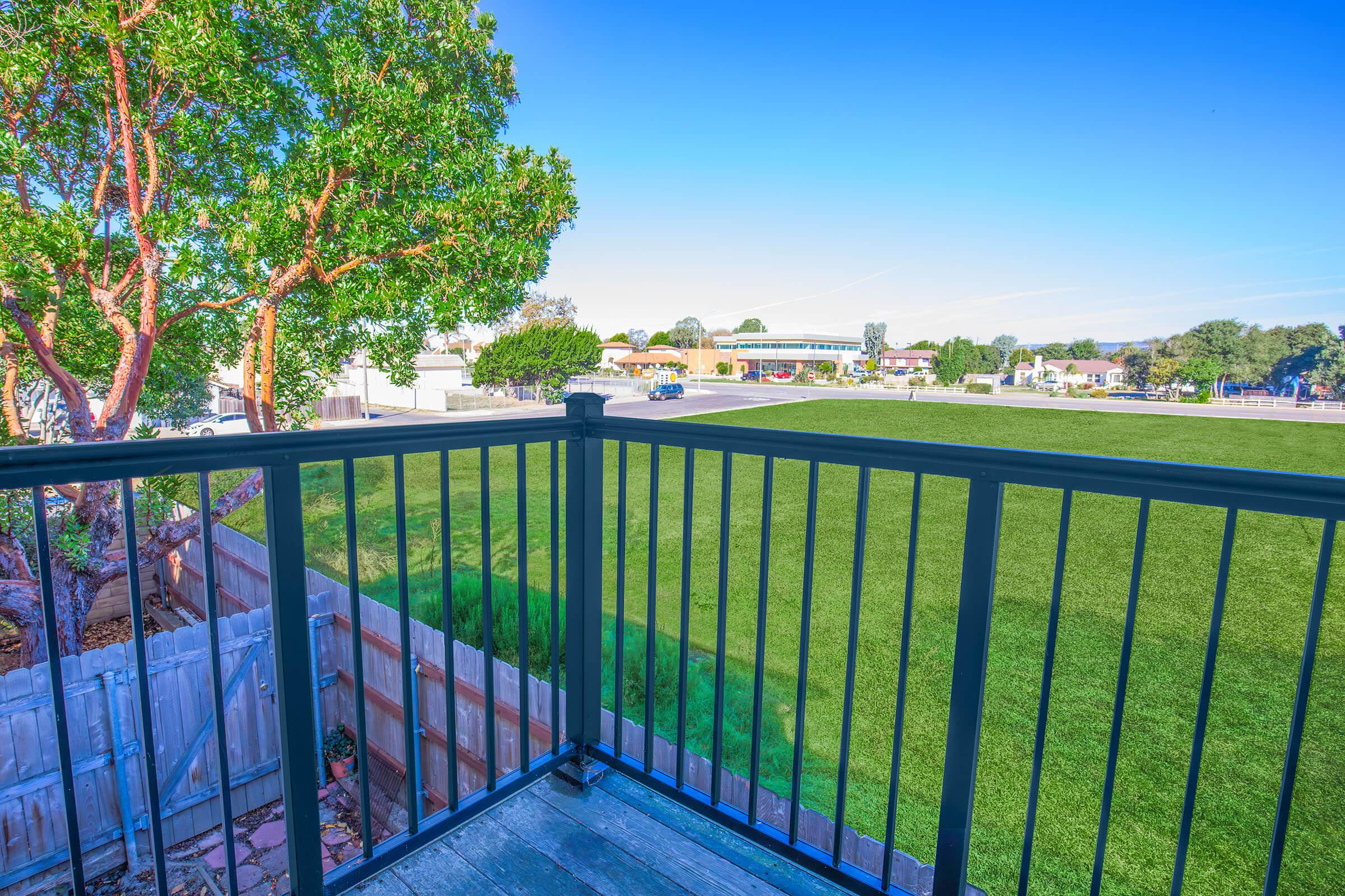 View from a balcony overlooking a green lawn and a small commercial area in the distance. The scene includes a clear blue sky and a nearby tree. The railing of the balcony is visible in the foreground, adding depth to the perspective.