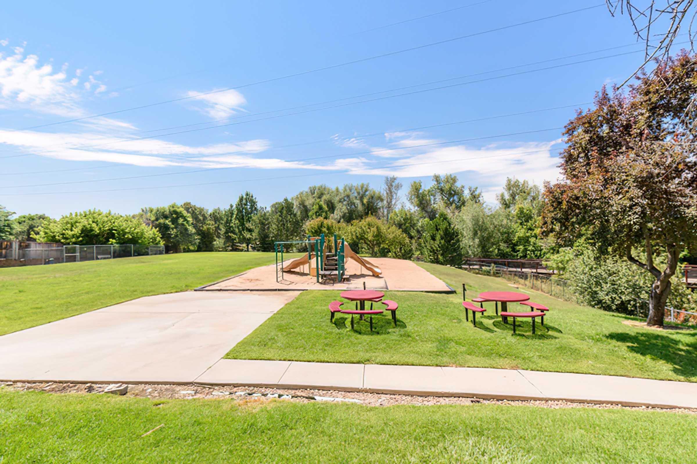 A sunny park scene featuring a playground with slides and climbing structures, surrounded by lush green grass and trees. Two red picnic tables are placed on a concrete path in the foreground. Blue skies with a few clouds complete the inviting outdoor setting.