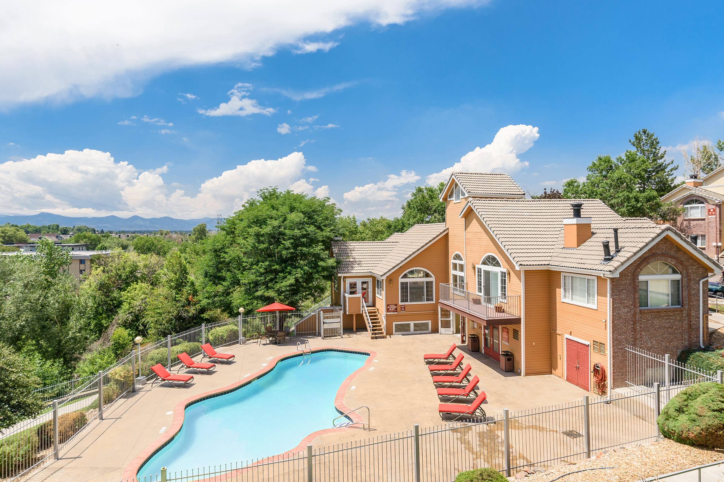 A bright, sunny outdoor swimming pool area featuring a light brown building with large windows, surrounded by greenery and trees. Red lounge chairs are neatly arranged around the pool, with an umbrella providing shade. The sky is clear with scattered clouds and distant mountains visible in the background.
