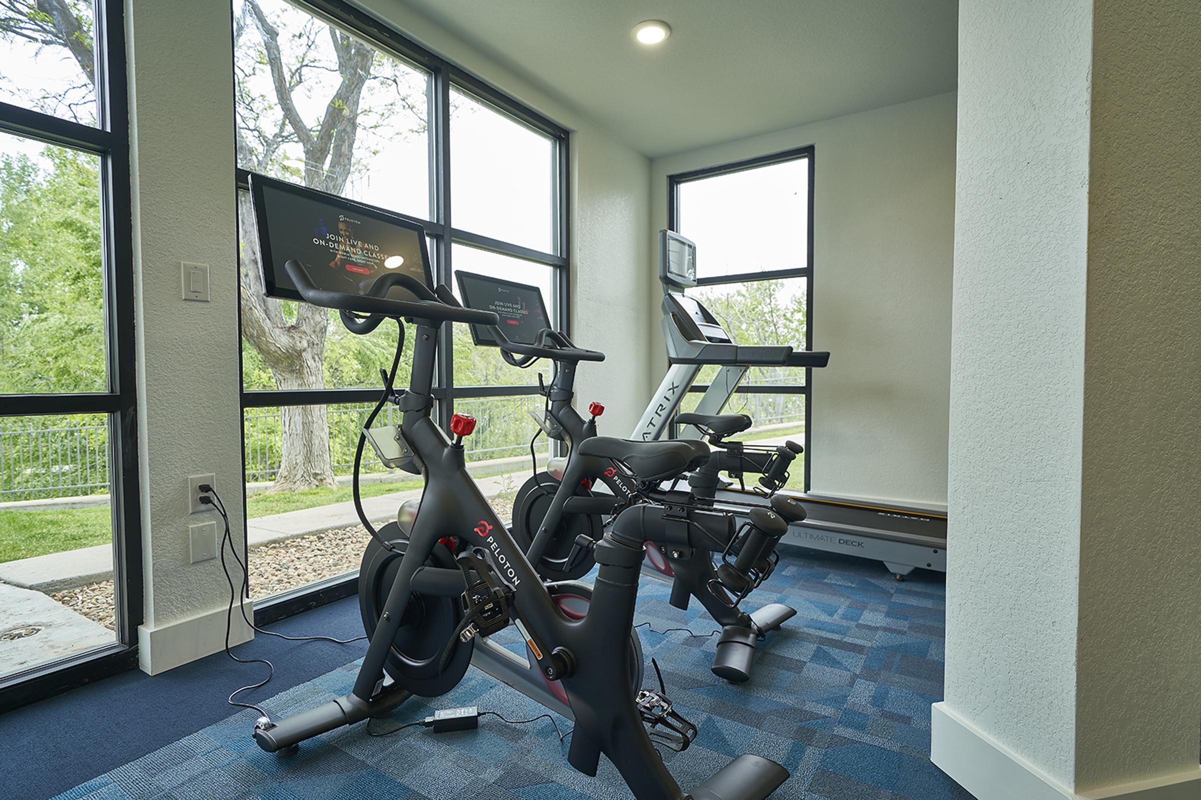 Indoor fitness area featuring two modern exercise bikes with screens, a treadmill in the background, large windows letting in natural light, and a view of greenery outside. The space is designed for workouts, with a clean and contemporary aesthetic.