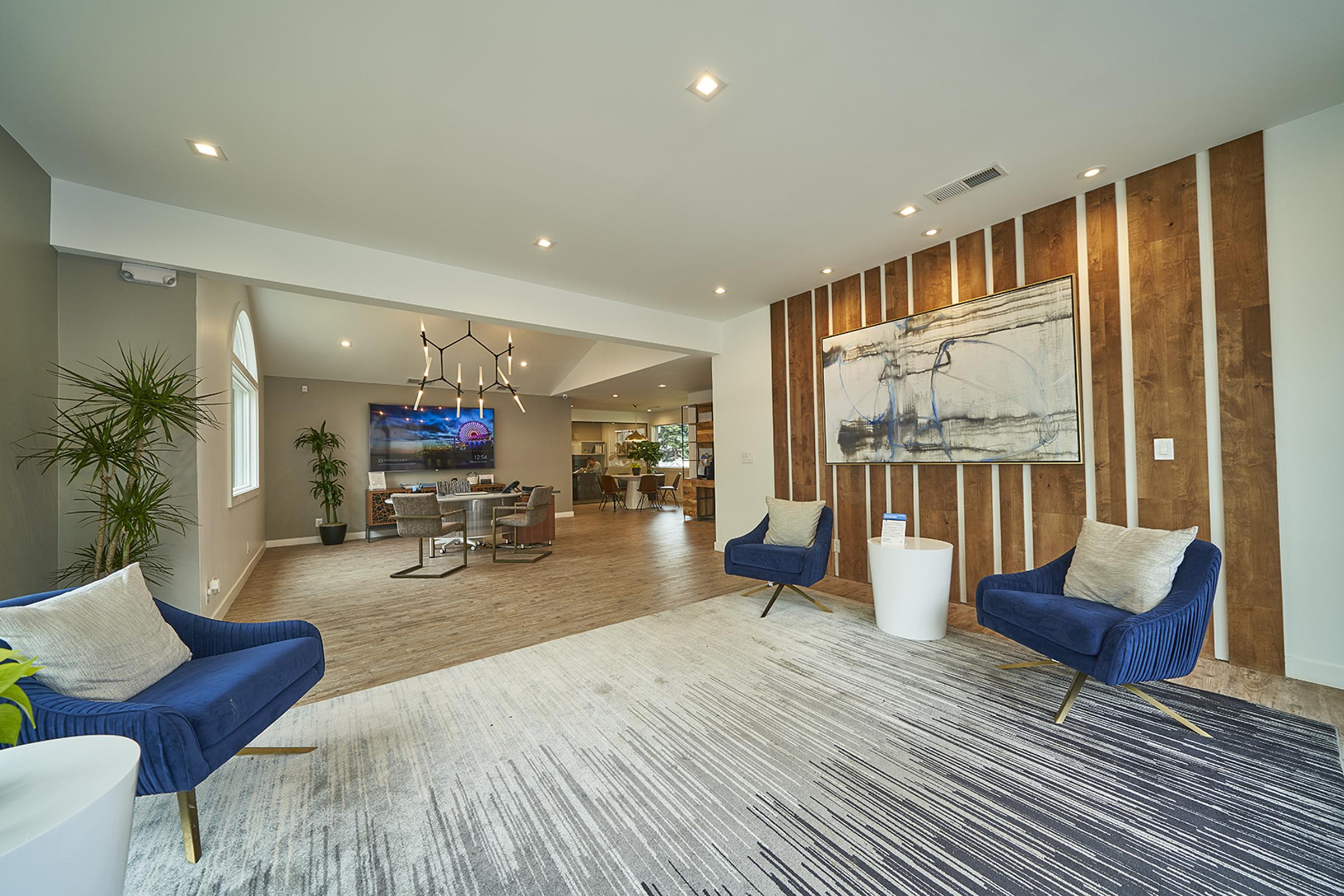 A modern living space featuring two blue accent chairs on a patterned rug, a stylish wooden accent wall with abstract art, and a view of a spacious open area with a TV and dining setup in the background. Potted plants add a touch of greenery to the decor.