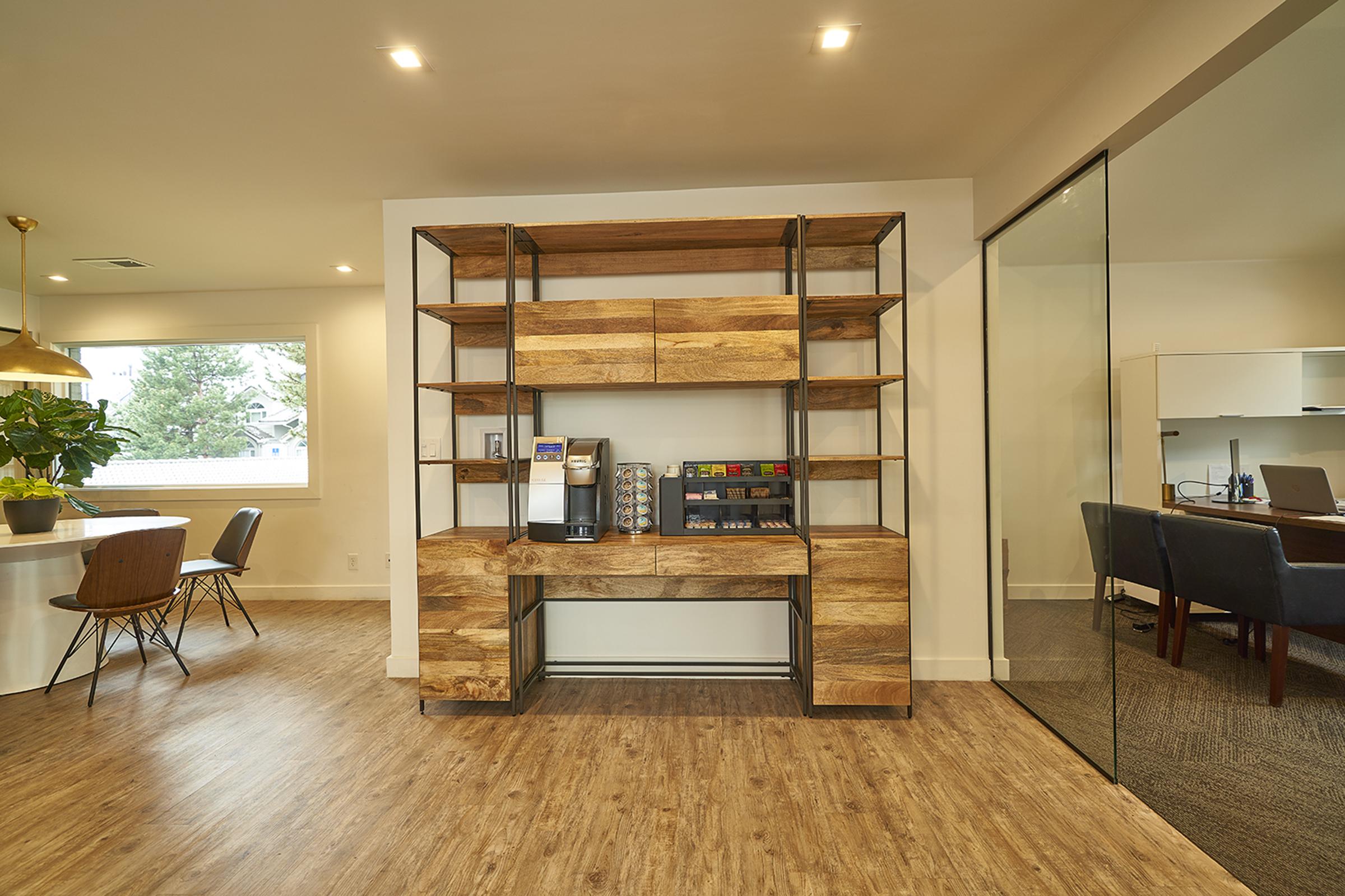 Modern office space featuring a wooden shelving unit with a coffee machine and colorful buttons. A round dining table with wooden chairs is visible, along with a glass-enclosed meeting room in the background. The decor is bright and welcoming, with natural light coming through the windows.
