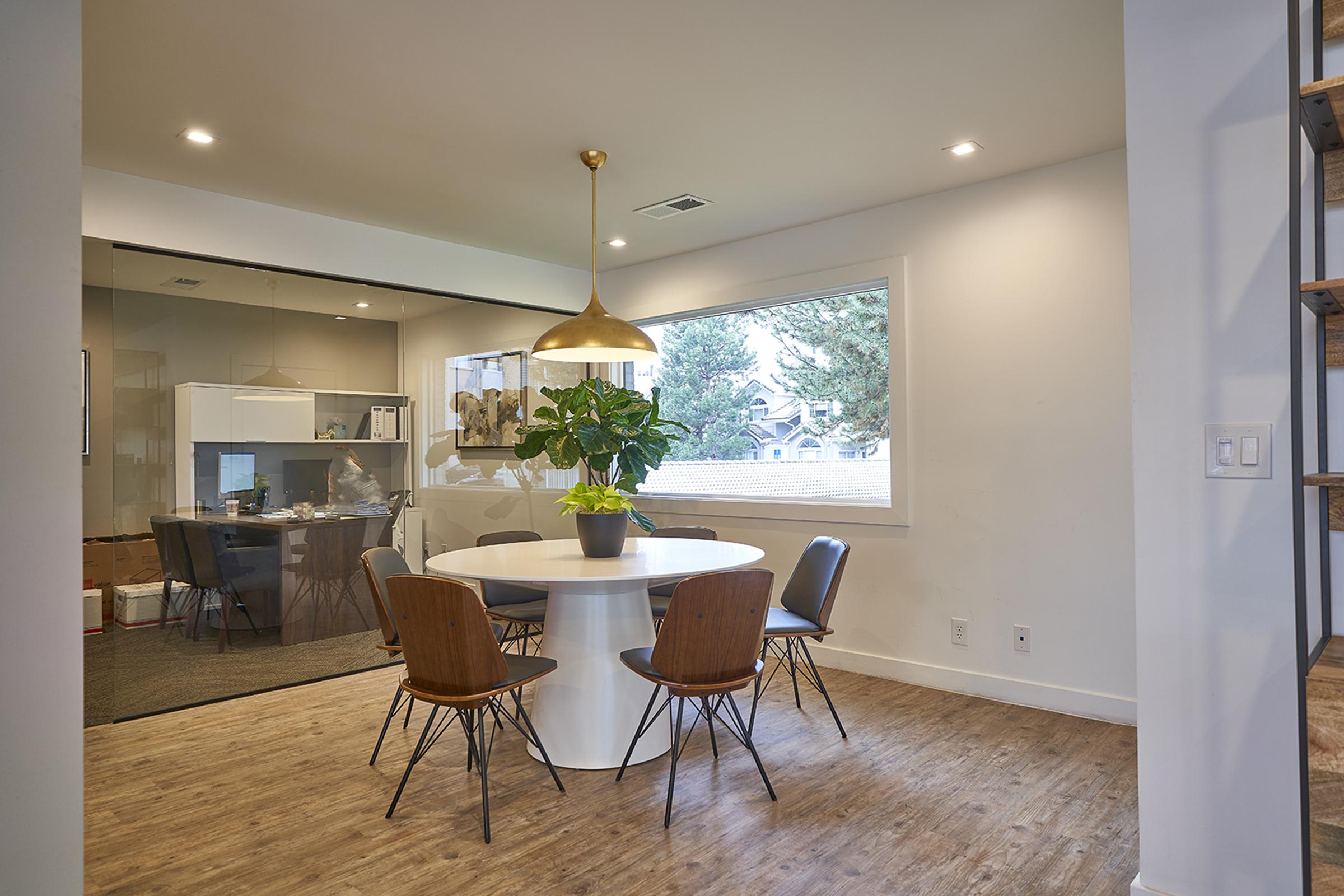 A modern dining area featuring a round white table surrounded by wooden chairs, a large potted plant as a centerpiece, and a pendant light hanging above. In the background, there is a glass-walled room with a table and chairs, and a window showcasing a view of trees outside.