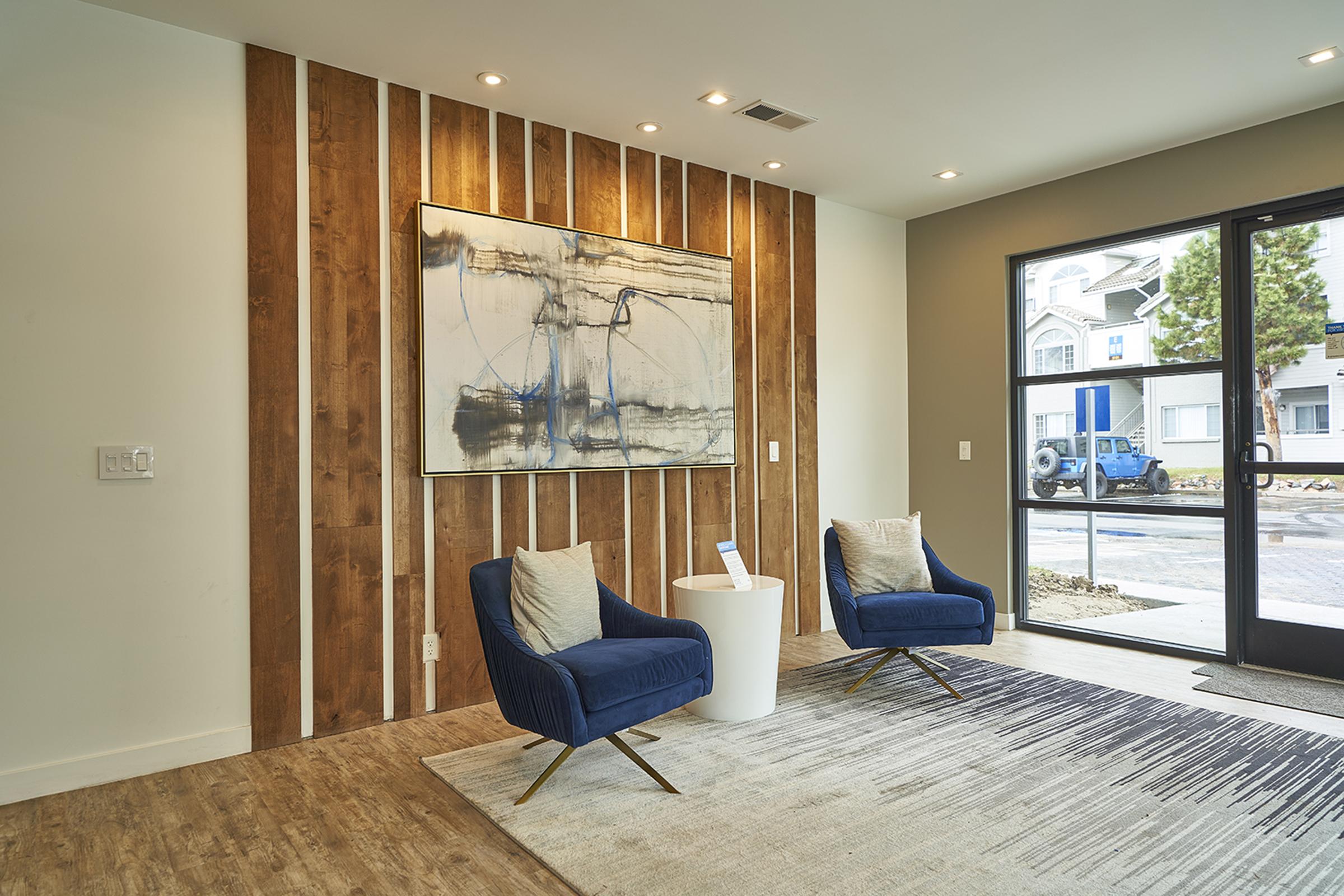 A modern interior space featuring two blue armchairs with cream cushions, placed on a patterned rug. Behind them is a wooden accent wall with vertical slats, and a large abstract painting above the chairs. A small white side table is positioned nearby, and large windows offer a view of the outside area.