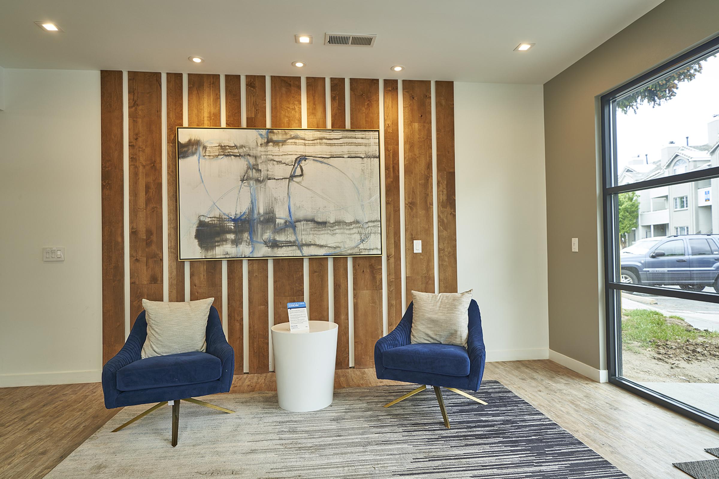 A modern interior space featuring two blue velvet chairs with cream pillows, a white circular table in between, and a large abstract painting on a wooden accent wall. Natural light enters through a large window, showcasing a view of the outside. The flooring is light wood, adding warmth to the design.