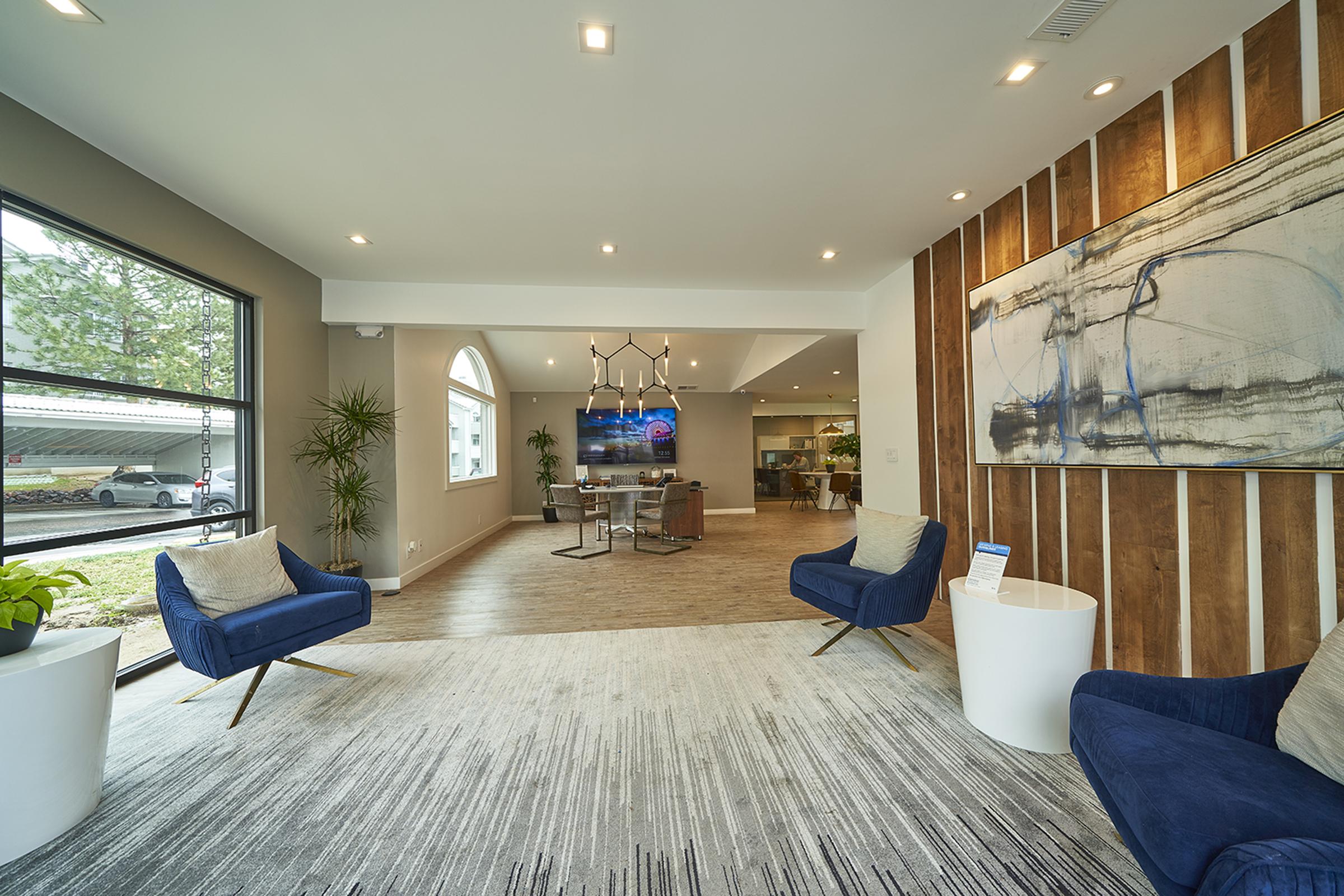 A modern interior lobby featuring two blue armchairs, a round white table, and a large abstract painting on a wooden accent wall. A television is mounted on the wall, and large windows provide natural light. Potted plants are placed throughout, creating a welcoming atmosphere.