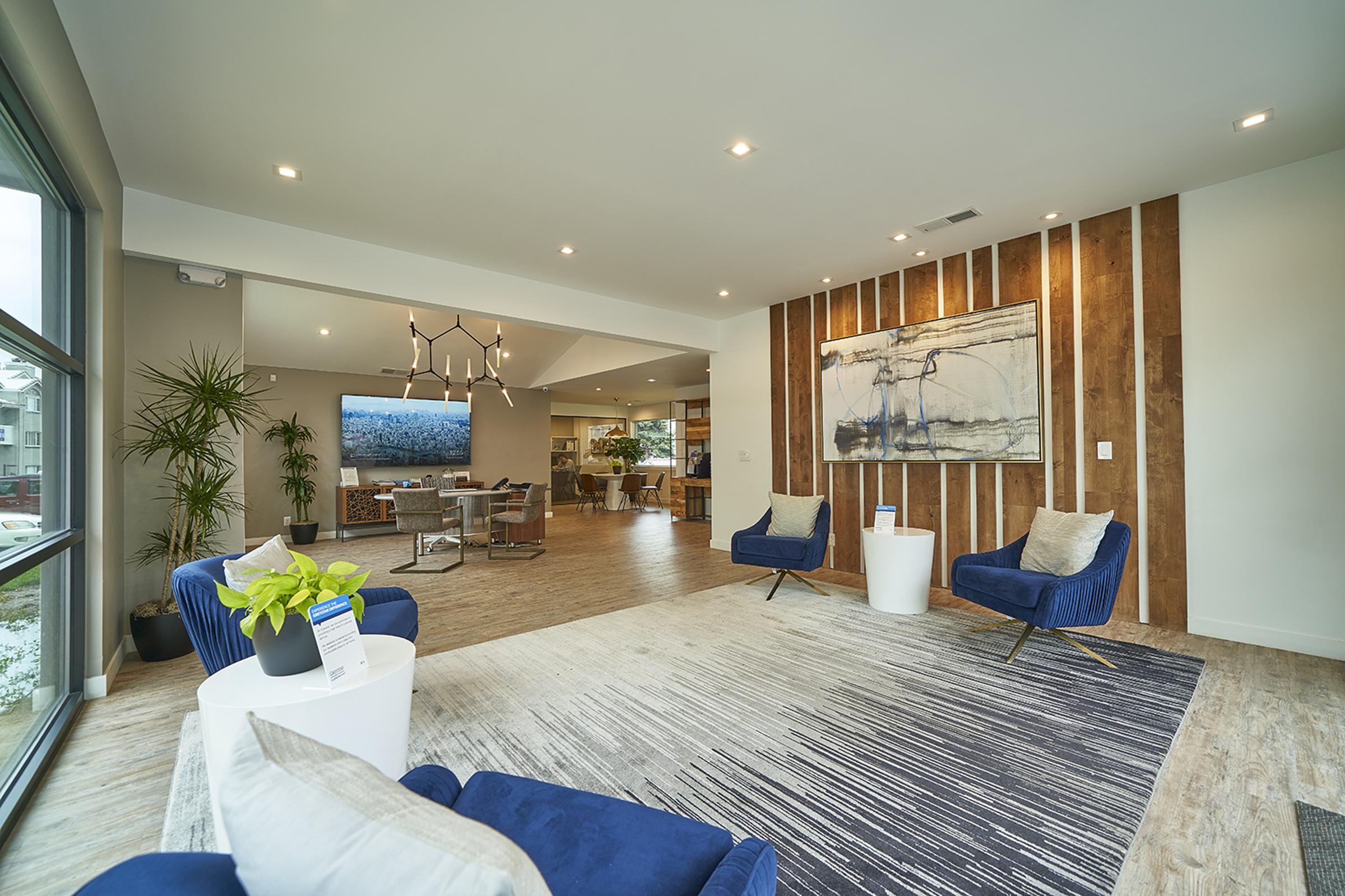 Modern open-concept living area with wood accent wall, featuring two blue armchairs and a white side table with a plant. A large abstract painting hangs on the wall, adjacent to a spacious lounge area with a television. Warm lighting enhances the inviting atmosphere of the stylish interior.