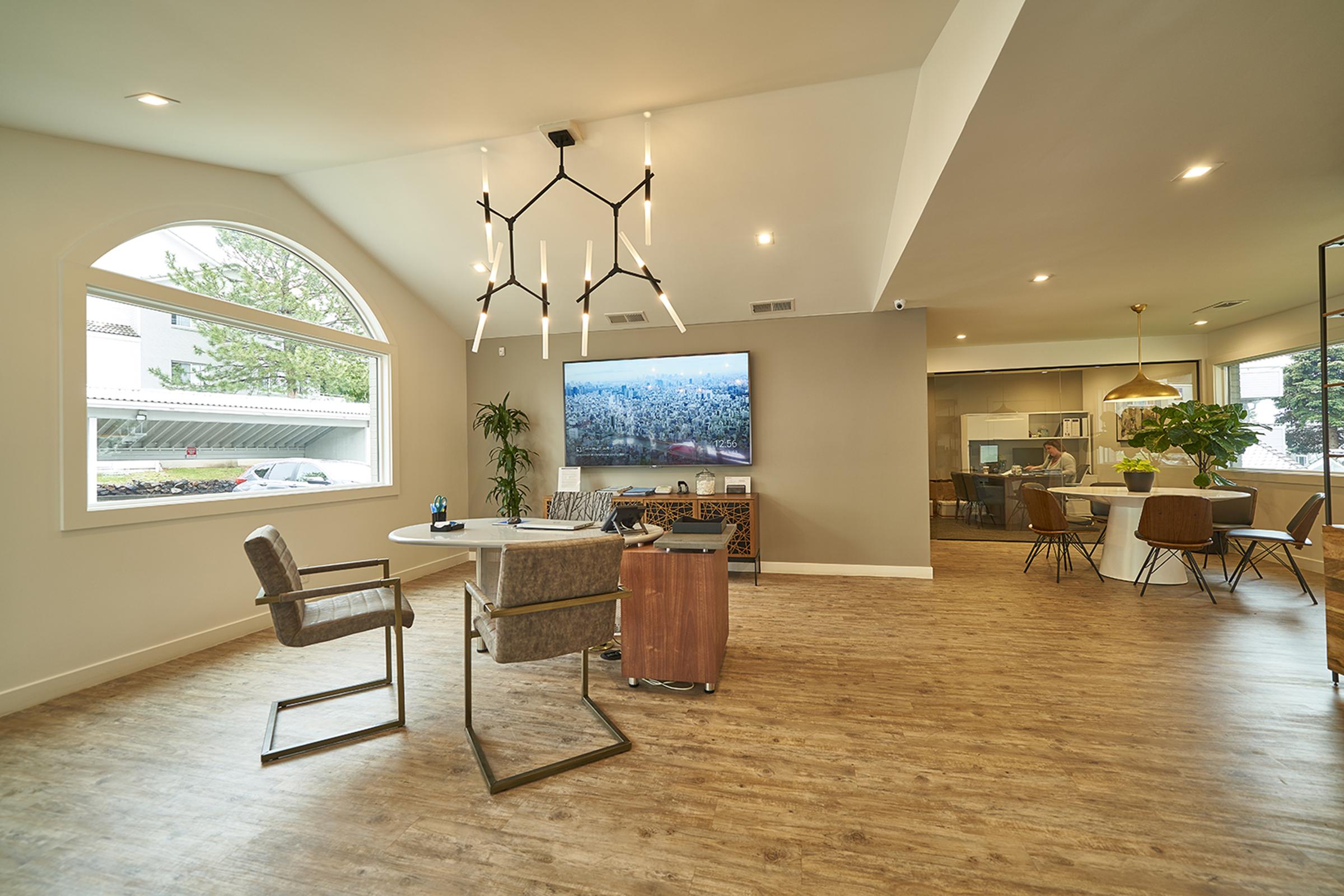 Modern living room featuring a stylish design with a large TV mounted on the wall, a round dining table, and two elegant chairs. A unique chandelier hangs from the ceiling, and large windows provide natural light. The space includes plants and different seating areas, creating an inviting atmosphere.