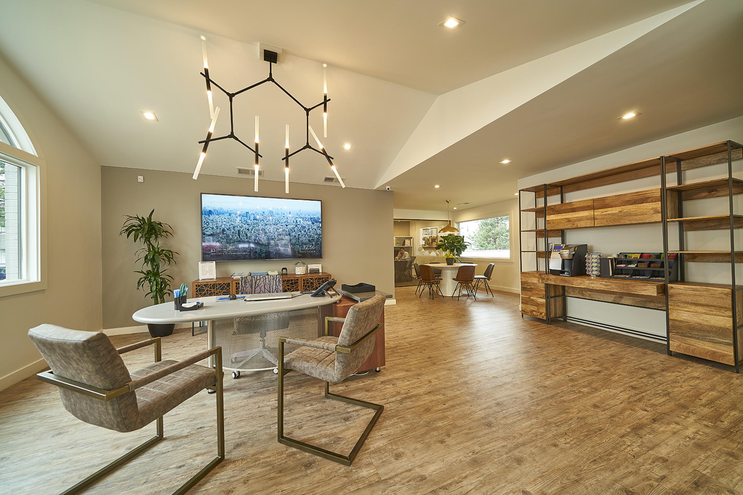 Modern interior of a spacious office or living area featuring a large TV mounted on the wall, a round table with two stylish chairs, a sleek bookshelf with decorative items, and large windows allowing natural light. The decor includes plants and contemporary lighting fixtures, creating a welcoming atmosphere.