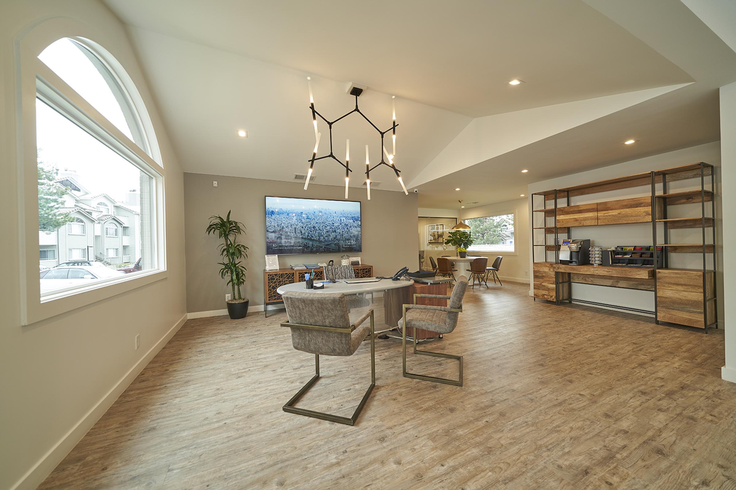 A modern, open-concept office space featuring a circular table with stylish chairs, a large wall-mounted screen displaying a cityscape, and a combination of wooden shelving and plants. Bright, natural light floods through large windows, enhancing the contemporary design.