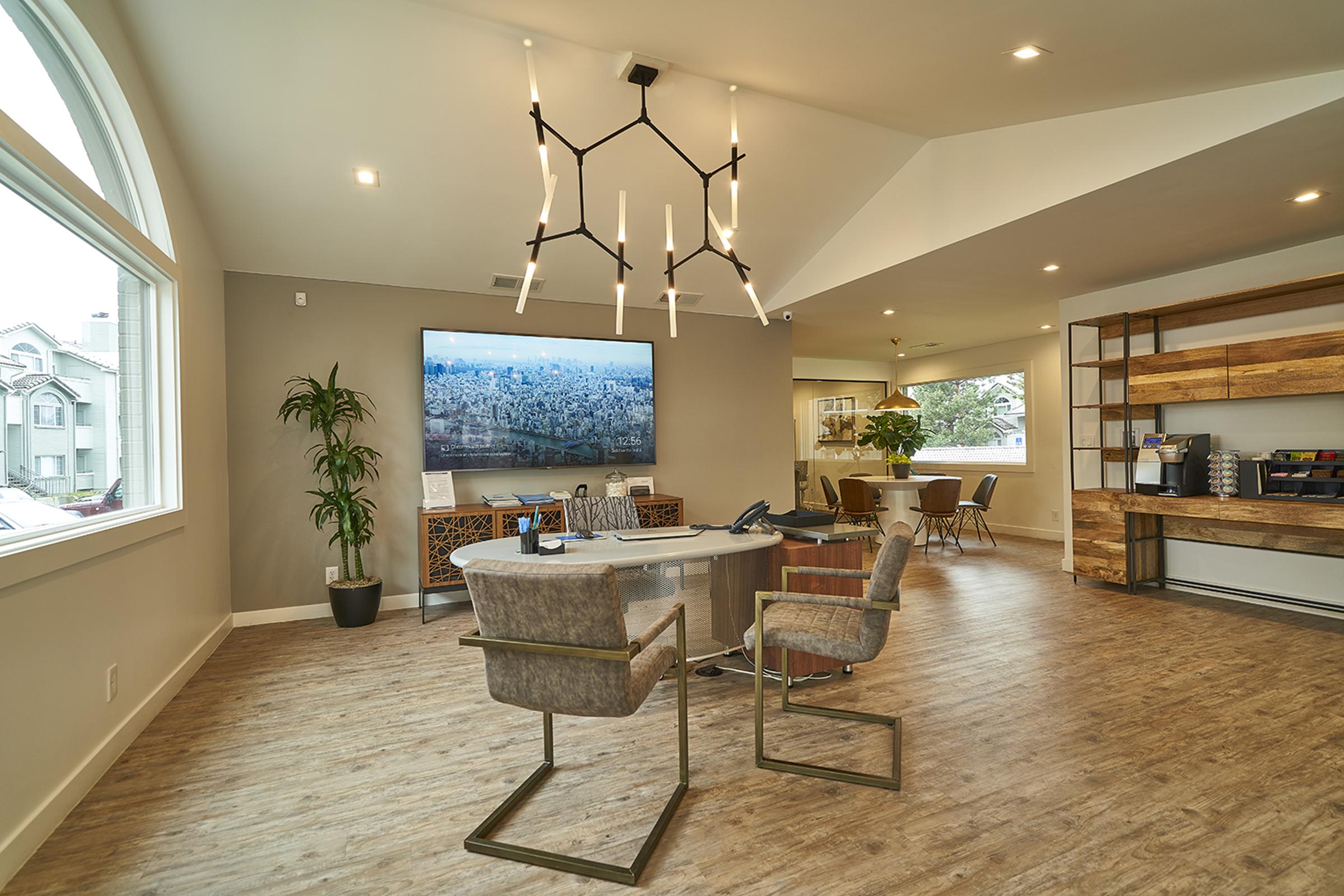 Modern office interior featuring a circular table with stylish chairs, a large wall-mounted TV displaying a scenic city view, a contemporary light fixture, and wooden shelving. A plant adds a touch of greenery to the space, which has neutral-colored walls and ample natural light from large windows.