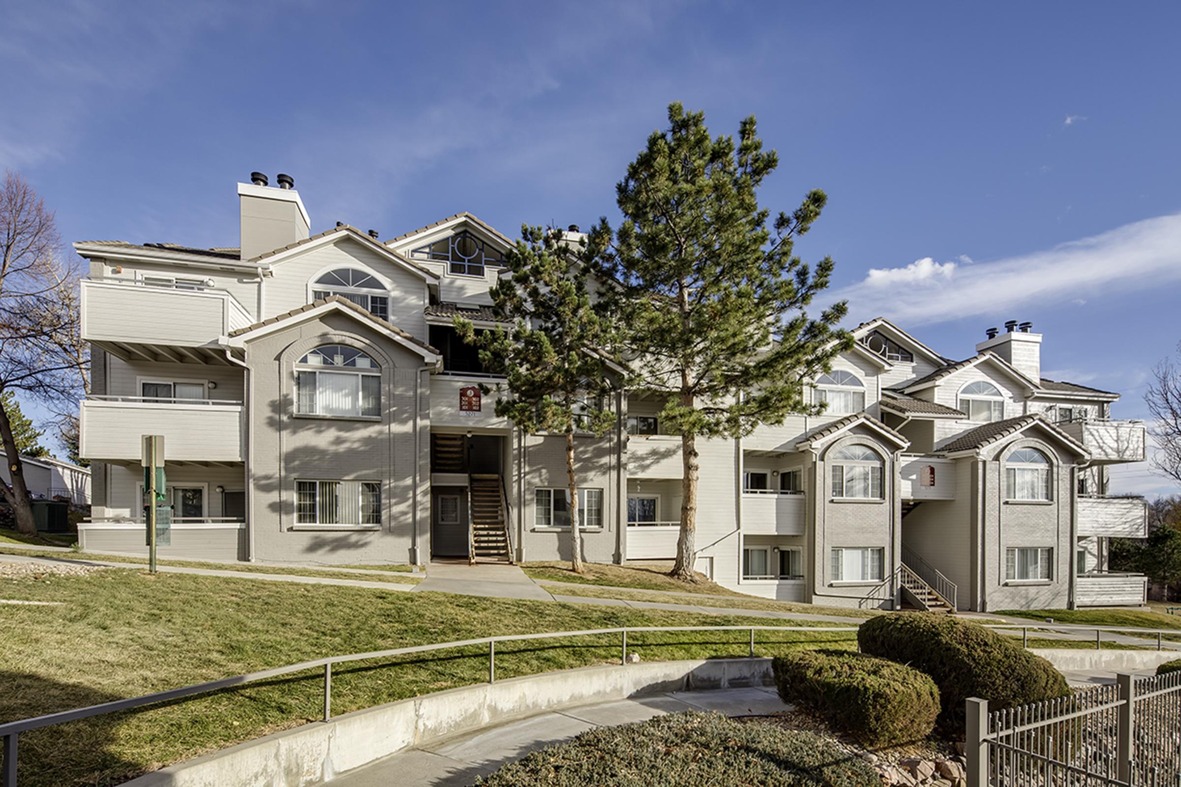 A multi-unit residential building featuring a modern architectural design. The structure has light gray siding, large windows, and a stairway leading to the entrance. Lush green grass and shrubs surround the building, with a clear blue sky in the background.