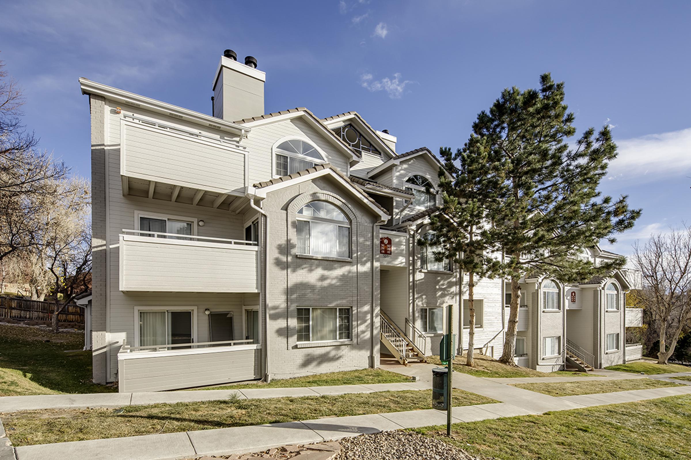 A multi-unit residential building with a light-colored exterior, featuring balconies and large windows. The building is surrounded by green grass, with a pathway leading to the entrance. Nearby, there are trees and a clear blue sky, indicating a pleasant day.