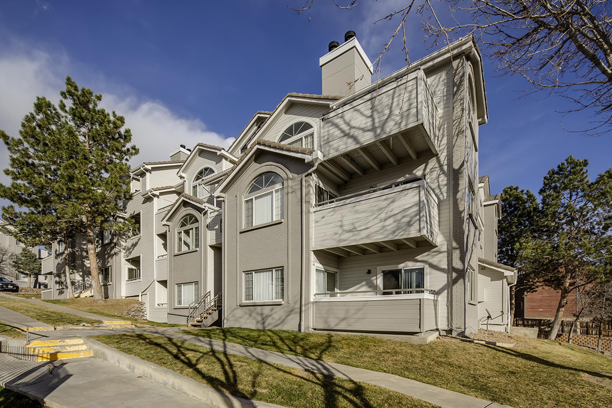 A multi-unit residential building with a light gray exterior, featuring multiple balconies and large windows. The architecture includes arched elements and a sloped roof. The surrounding area has grassy lawns and trees, with a clear blue sky in the background.