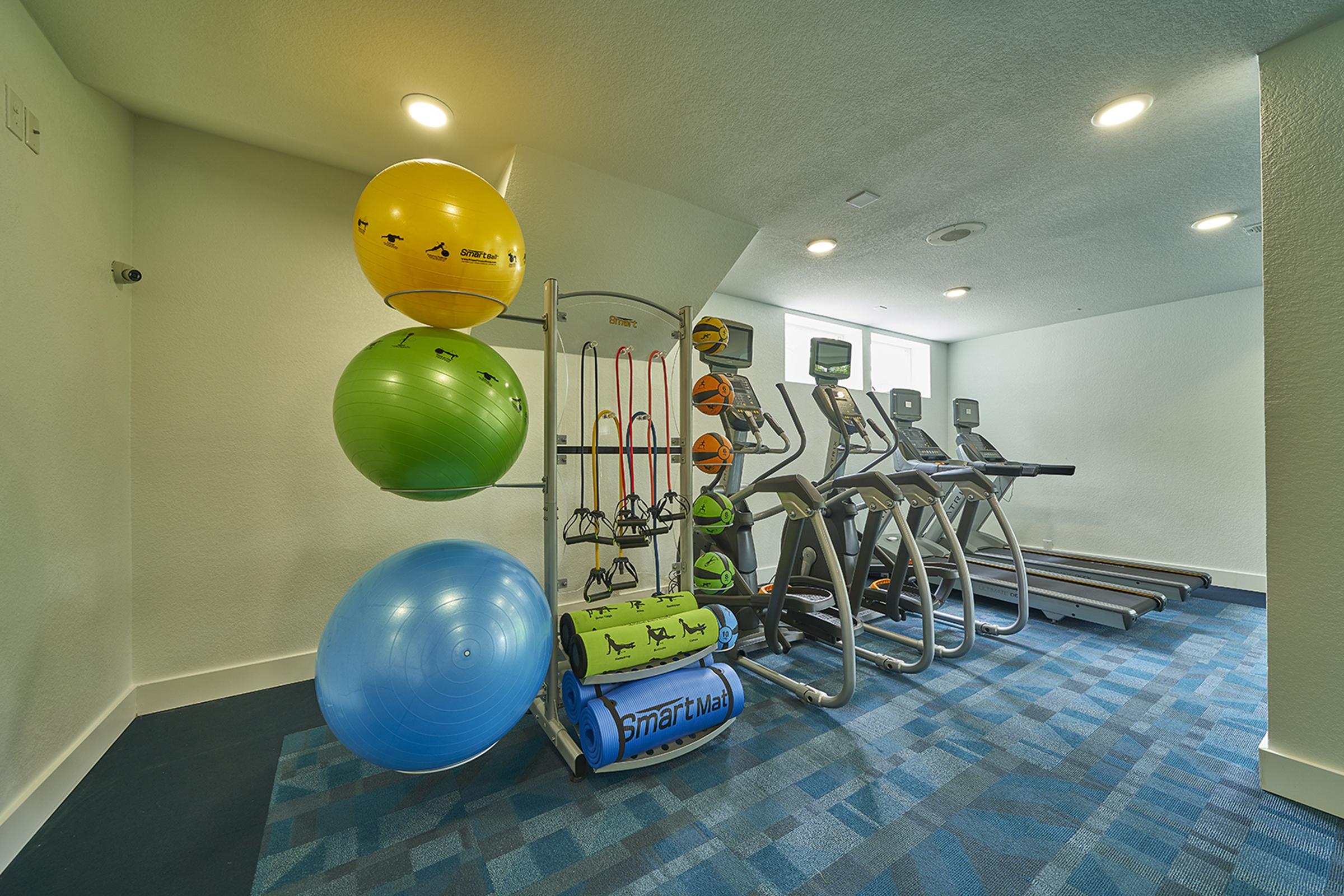 A well-equipped gym area featuring exercise equipment such as treadmills and stationary bikes. Colorful exercise balls are stacked, and there are resistance bands and weights on a rack, all set against a light-colored wall and carpeted blue flooring.