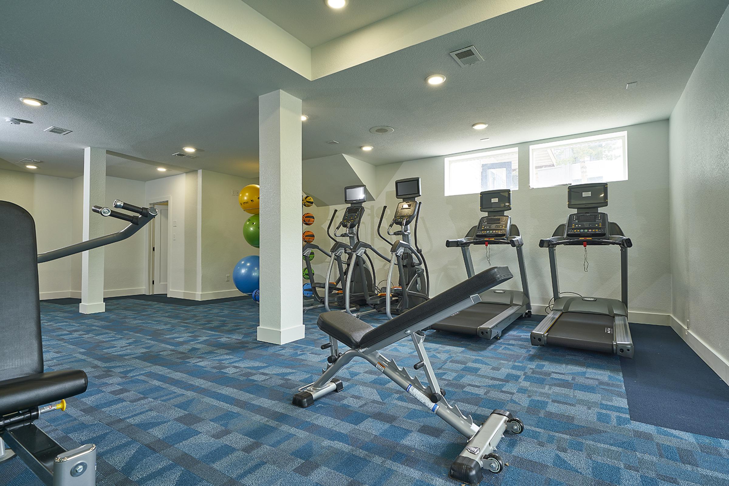 A modern gym interior featuring exercise equipment such as treadmills and elliptical machines, a weight bench, and exercise balls in various colors. The space has a clean, well-lit design with blue carpeting and white walls, creating a welcoming atmosphere for workouts.