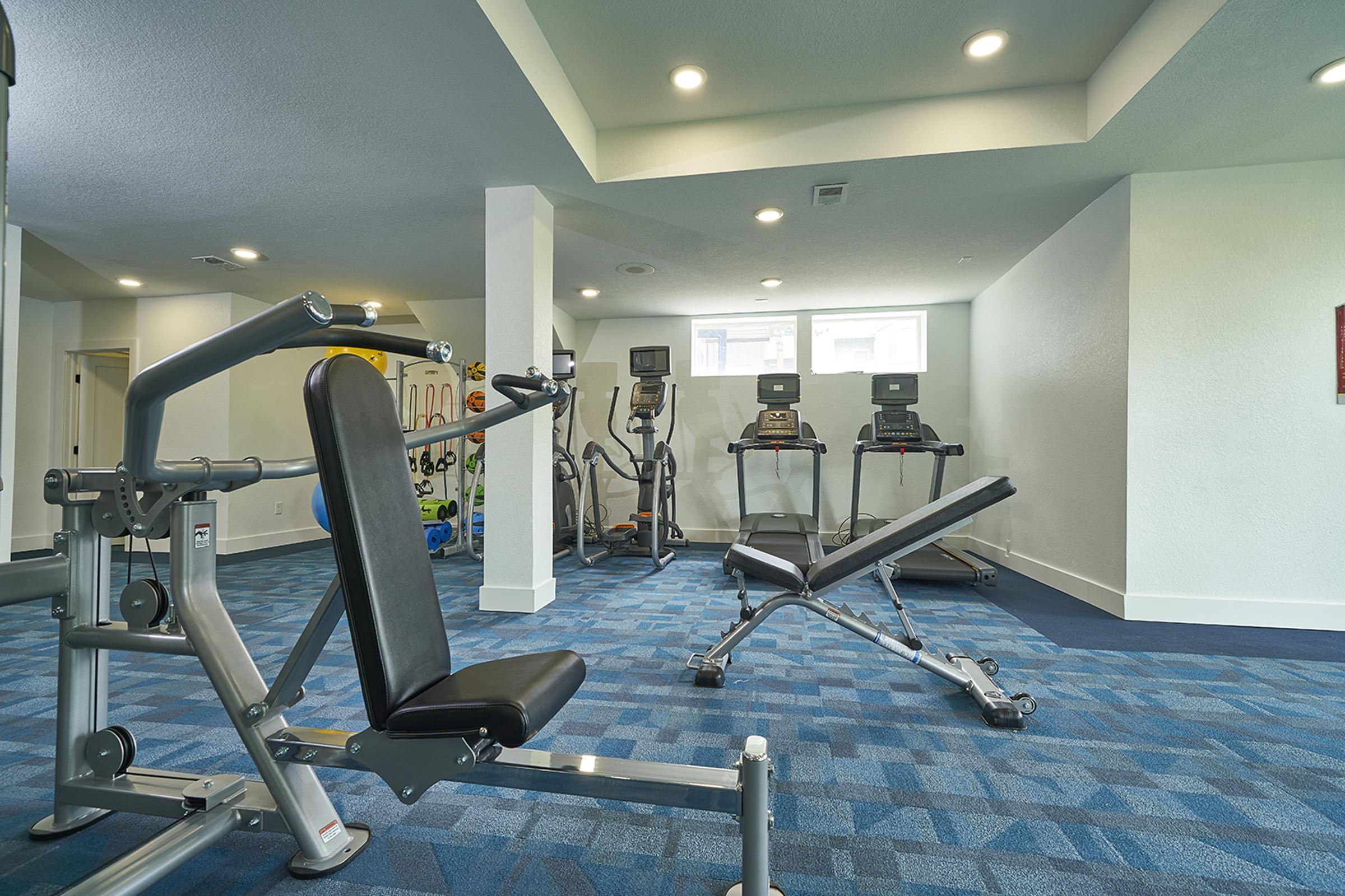 A modern gym interior featuring exercise equipment. In the foreground, a weight bench and a strength training machine are visible. In the background, several cardio machines like treadmills and ellipticals are lined up. The floor is carpeted in blue tones, and the space has bright, even lighting.