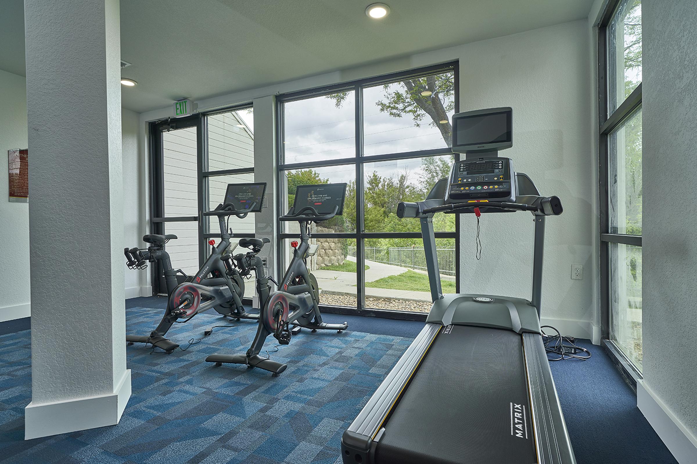 A modern gym area featuring equipment including two stationary bikes and a treadmill, all placed beside large windows offering a view of the outdoors. The space has bright lighting and a clean, carpeted floor, creating an inviting atmosphere for exercise.