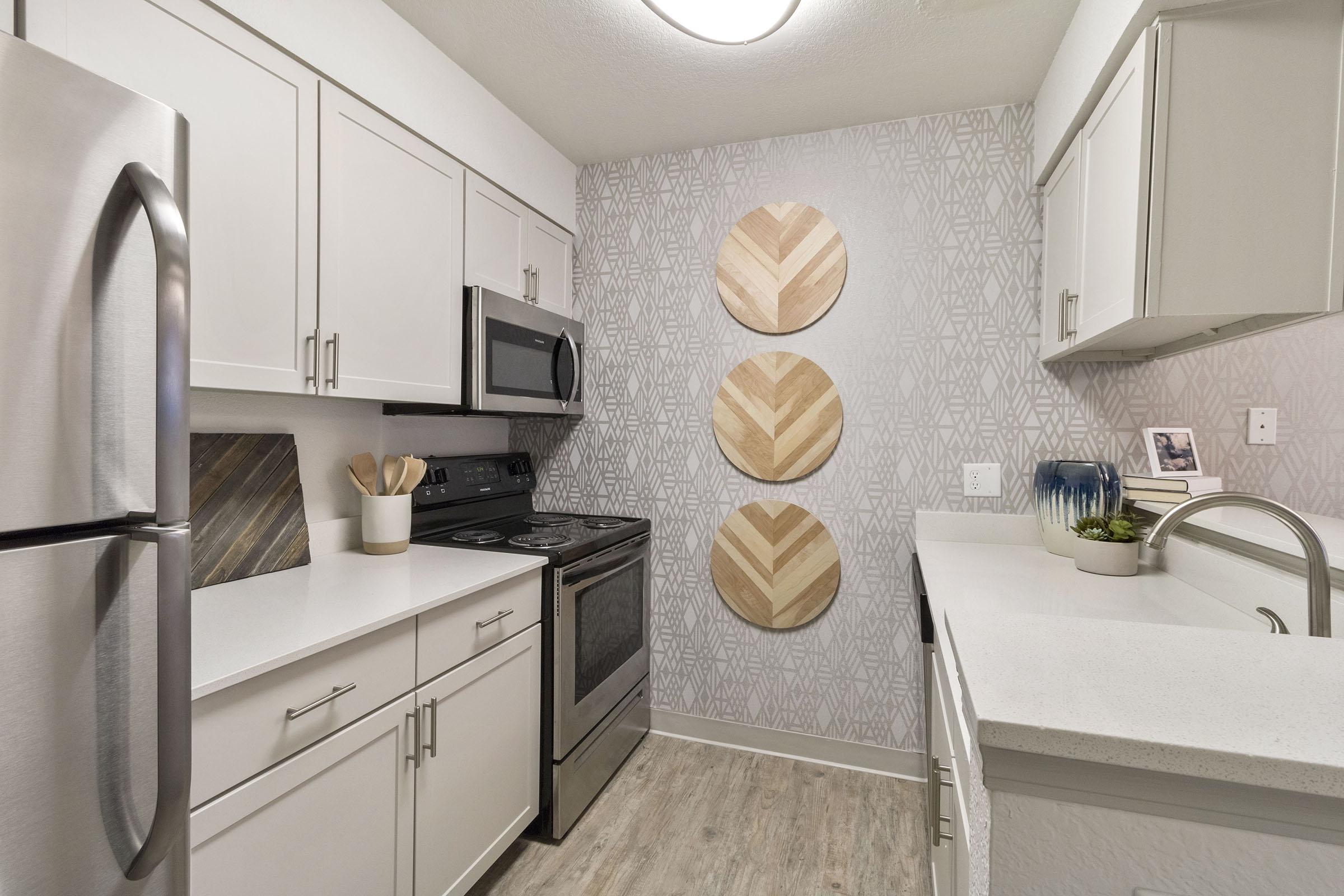 A modern kitchen featuring white cabinets, a stainless steel refrigerator and oven, and a gray geometric-patterned wall. Three decorative wooden wall pieces are mounted on one wall. The countertop is white with a small plant decoration, and there's a sink with a faucet.