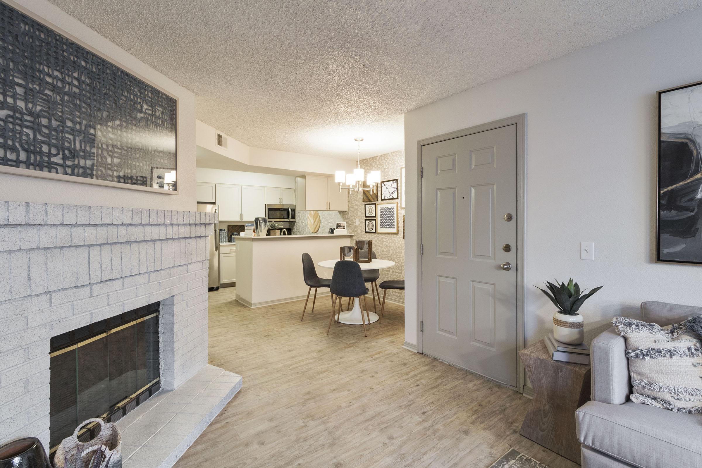 A spacious living area with a fireplace, light-colored walls, and a textured ceiling. A dining table with four chairs is visible, along with a kitchen area in the background. Decorative elements, including artwork and a potted plant, add warmth to the modern aesthetic of the room.