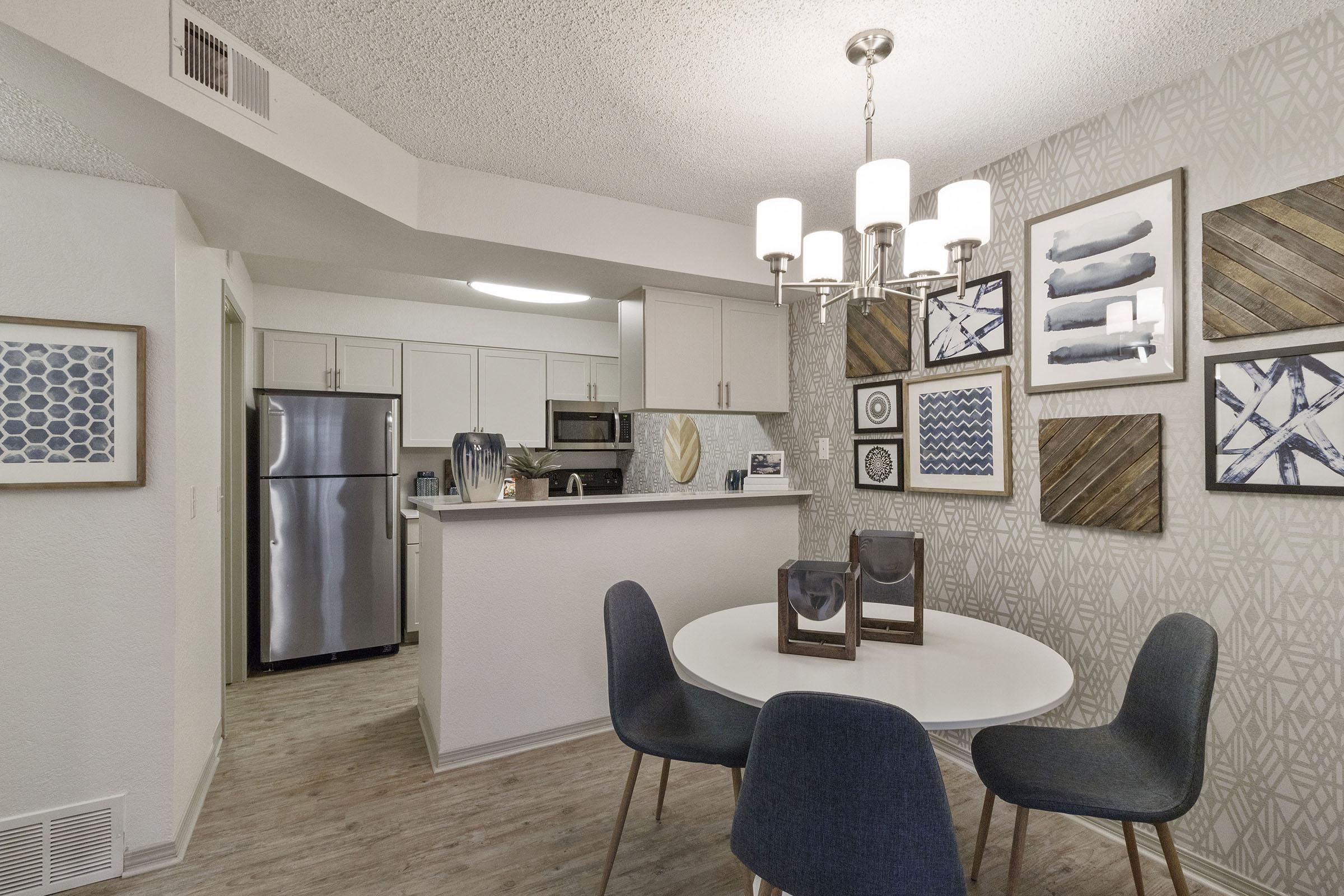 A modern dining area featuring a round white table surrounded by dark blue chairs, with a stylish chandelier above. In the background, there's a kitchen with stainless steel appliances and a unique wall decorated with framed artwork in blue and white tones, complementing the neutral color scheme.