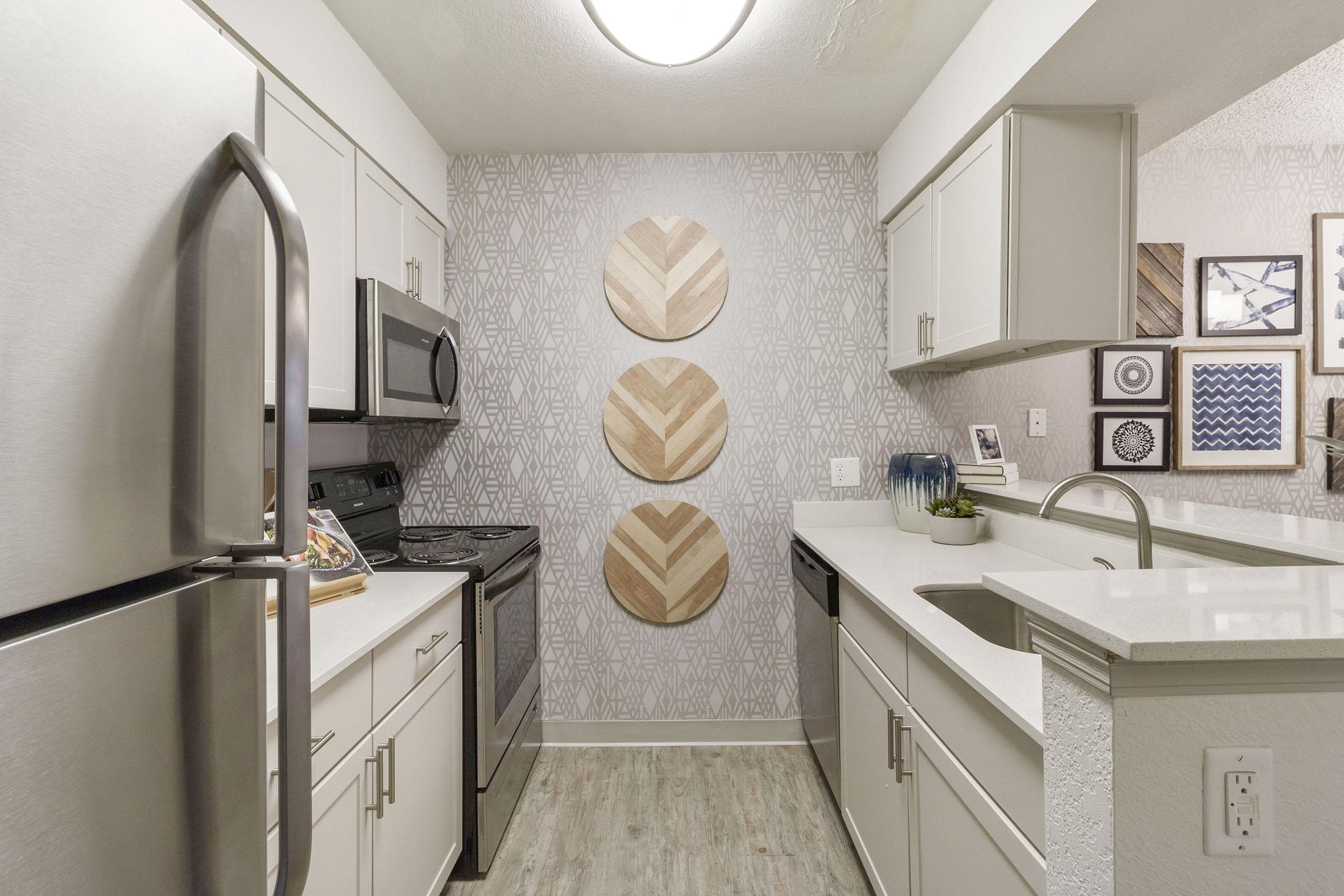 A modern kitchen featuring stainless steel appliances, including a refrigerator and stove. The cabinetry is light gray, and the counter has a sleek white surface. A decorative wall with three patterned wooden circles adds interest, while framed artwork and a plant accentuate the space. Bright lighting illuminates the area.