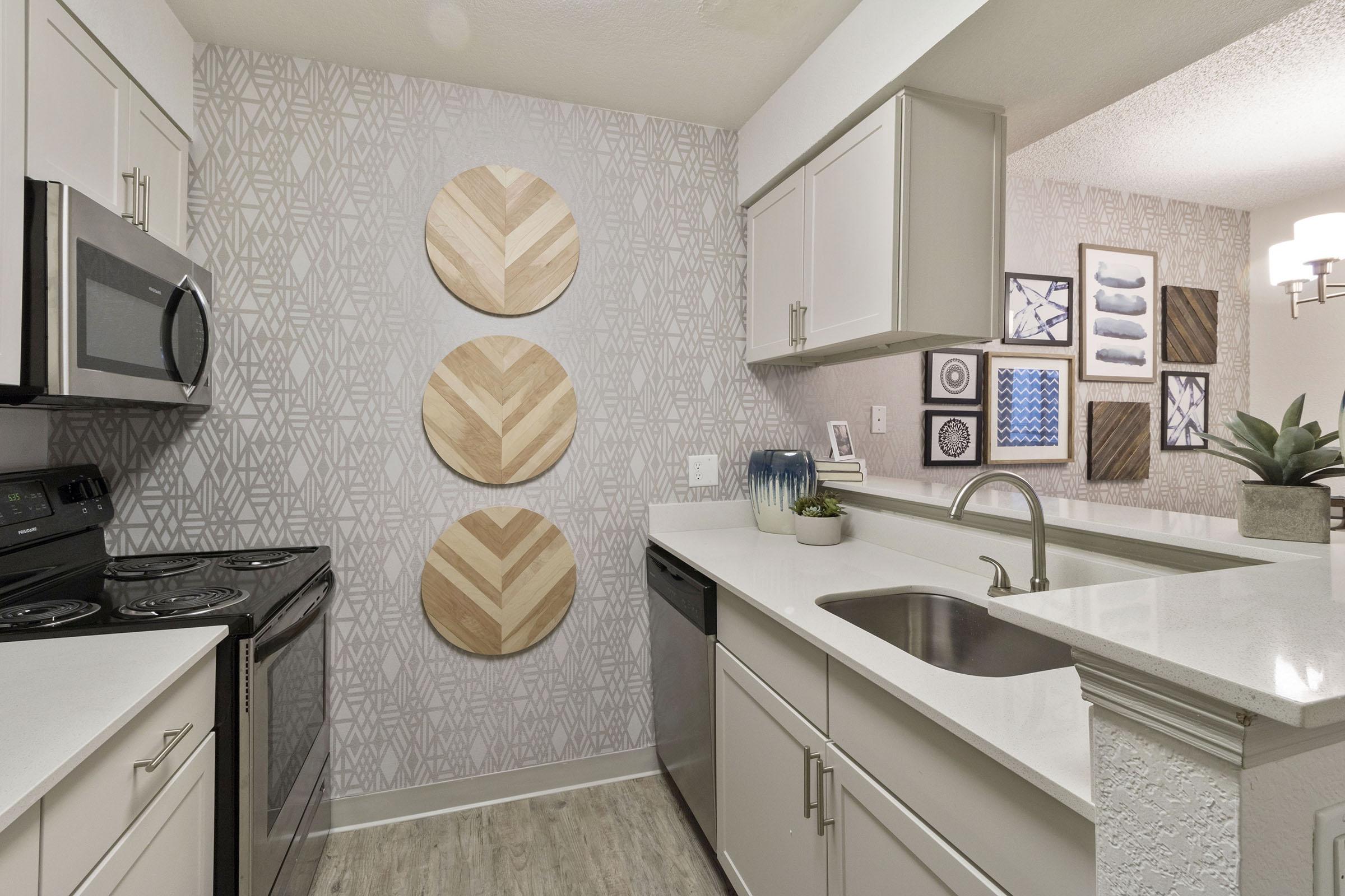 A modern kitchen featuring light-colored cabinetry, stainless steel appliances, and a decorative backsplash. The walls have a geometric pattern, and three wooden circular wall hangings add visual interest. A stylish plant is placed on the countertop, complementing the contemporary decor.