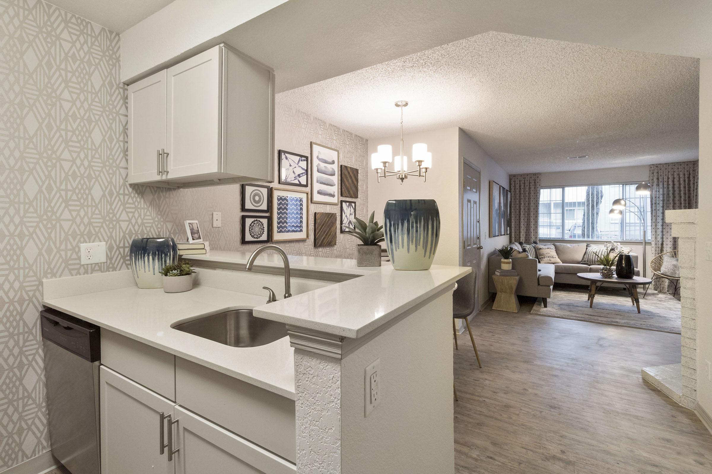 Modern kitchen view featuring a sleek sink and countertop, with a stylish backsplash. In the background, a cozy living area with a seating arrangement, decorative plants, and wall art creating a warm atmosphere. Natural light filters through window treatments. Elegant decor enhances the inviting ambiance.