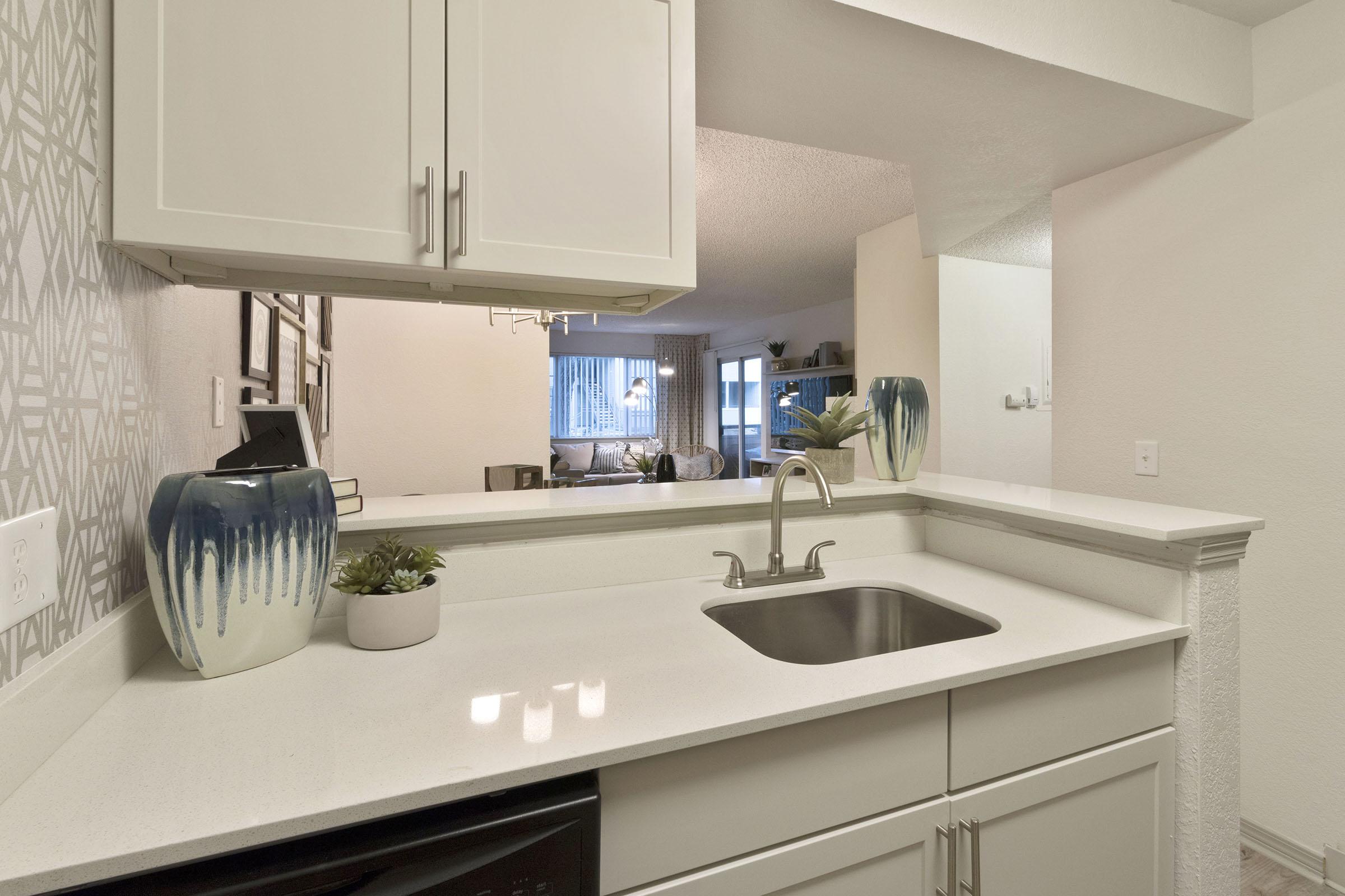 A modern kitchen with white cabinets and a sleek countertop featuring a sink. Decorative vases and a small plant add a touch of greenery. The background shows a living area with large windows and light-colored furnishings, creating a bright and inviting atmosphere.