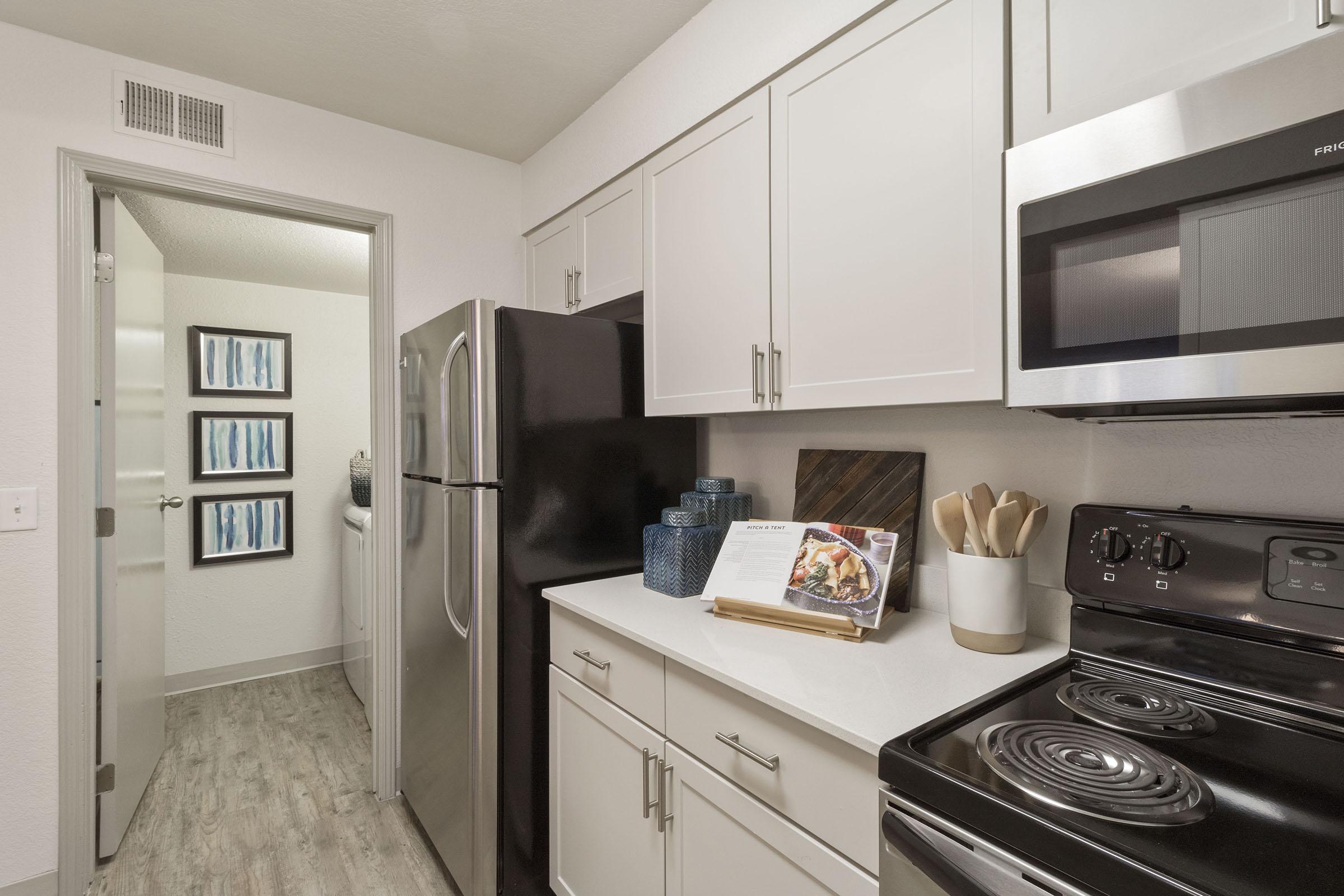 A modern kitchen featuring white cabinets, a black refrigerator, and a stainless steel oven and microwave. The countertop is tidy, with decorative items including a cookbook on a stand. The room has a light hardwood floor, and a doorway leads to another area, while two framed artworks hang on the wall.