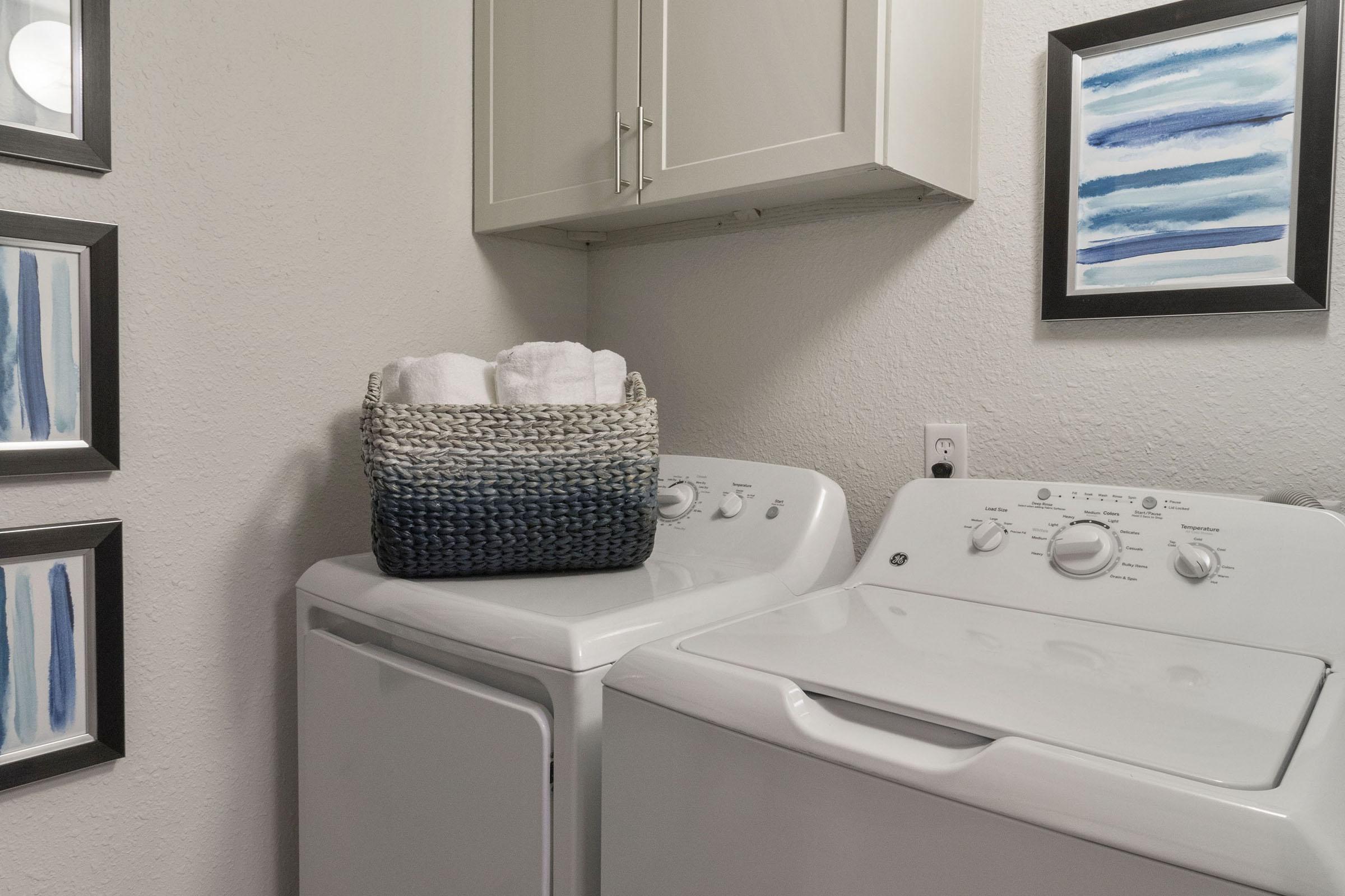 A laundry room featuring a white washing machine and dryer. There is a woven basket with towels on top of the dryer. The walls are adorned with framed artwork displaying blue and white abstract stripes, and there are cabinets above the appliances. The overall atmosphere is clean and organized.