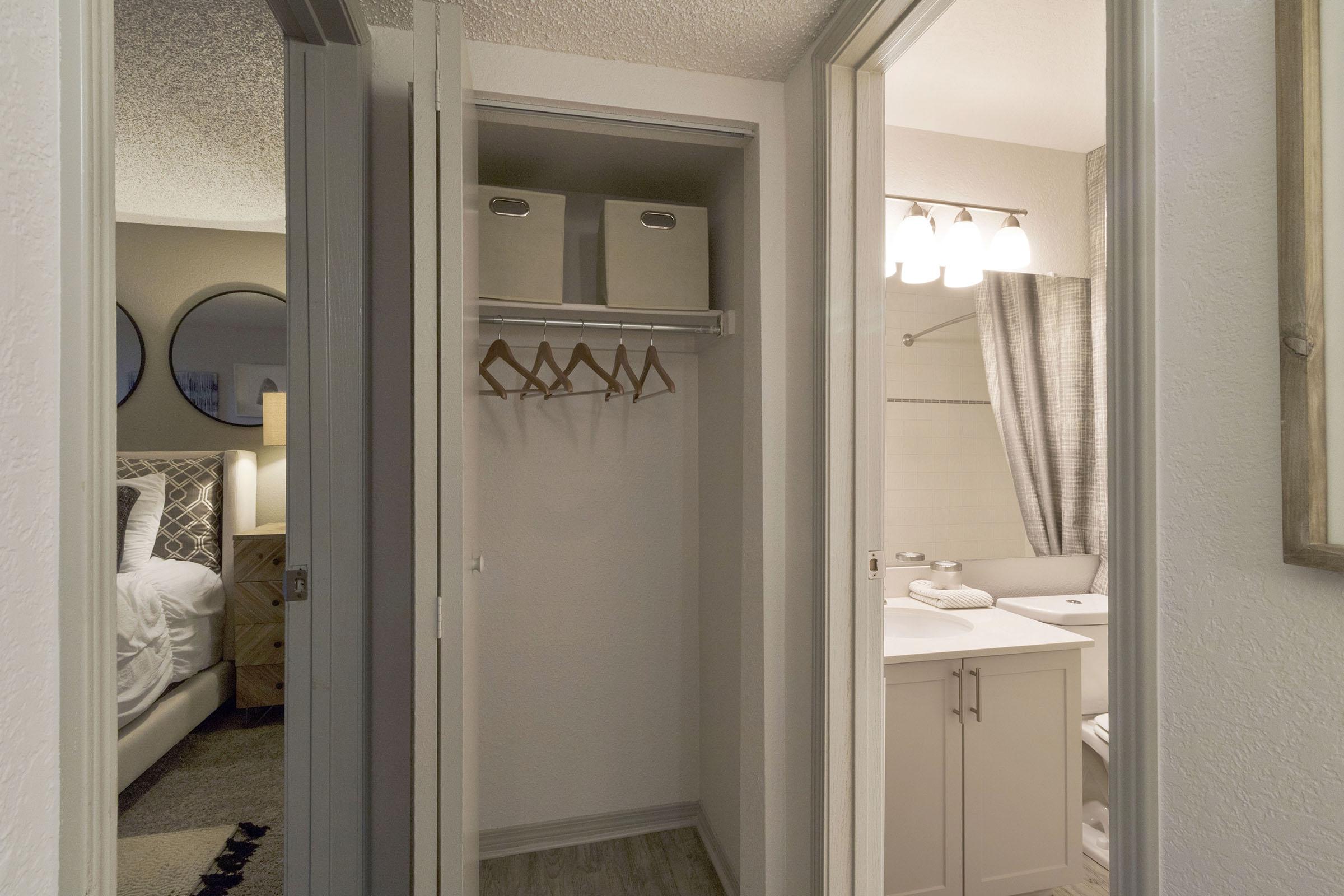 A view of a hotel room entrance with a closet on the left containing hangers and storage boxes. Beyond the closet, a bathroom can be seen with a white vanity and shower. The room's decor includes circular mirrors and a neatly made bed visible in the background, contributing to a cozy atmosphere.