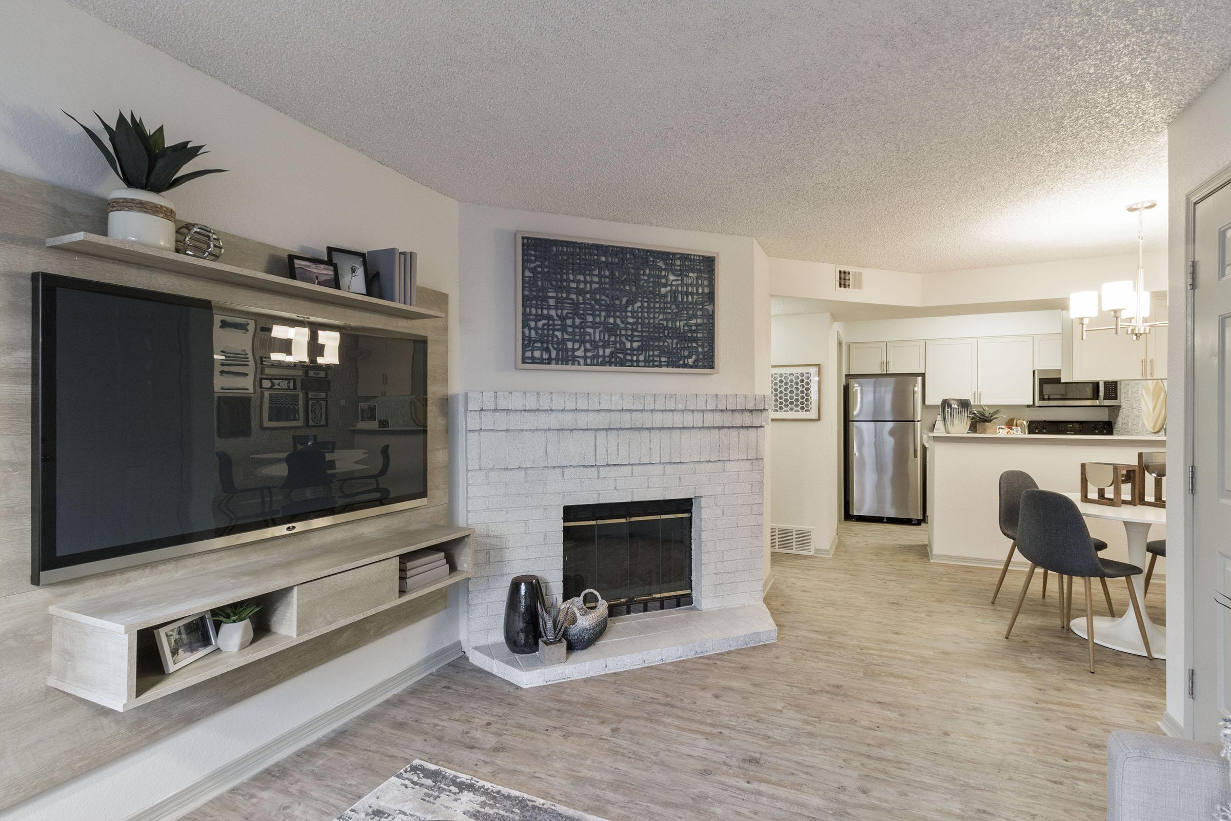 Living room featuring a mounted flat-screen TV above a light-colored fireplace. There is a wooden shelf with decor items on the left. A dining area is visible in the background, with a modern kitchen, a dining table, and gray chairs. The interior design has neutral tones and contemporary furnishings.
