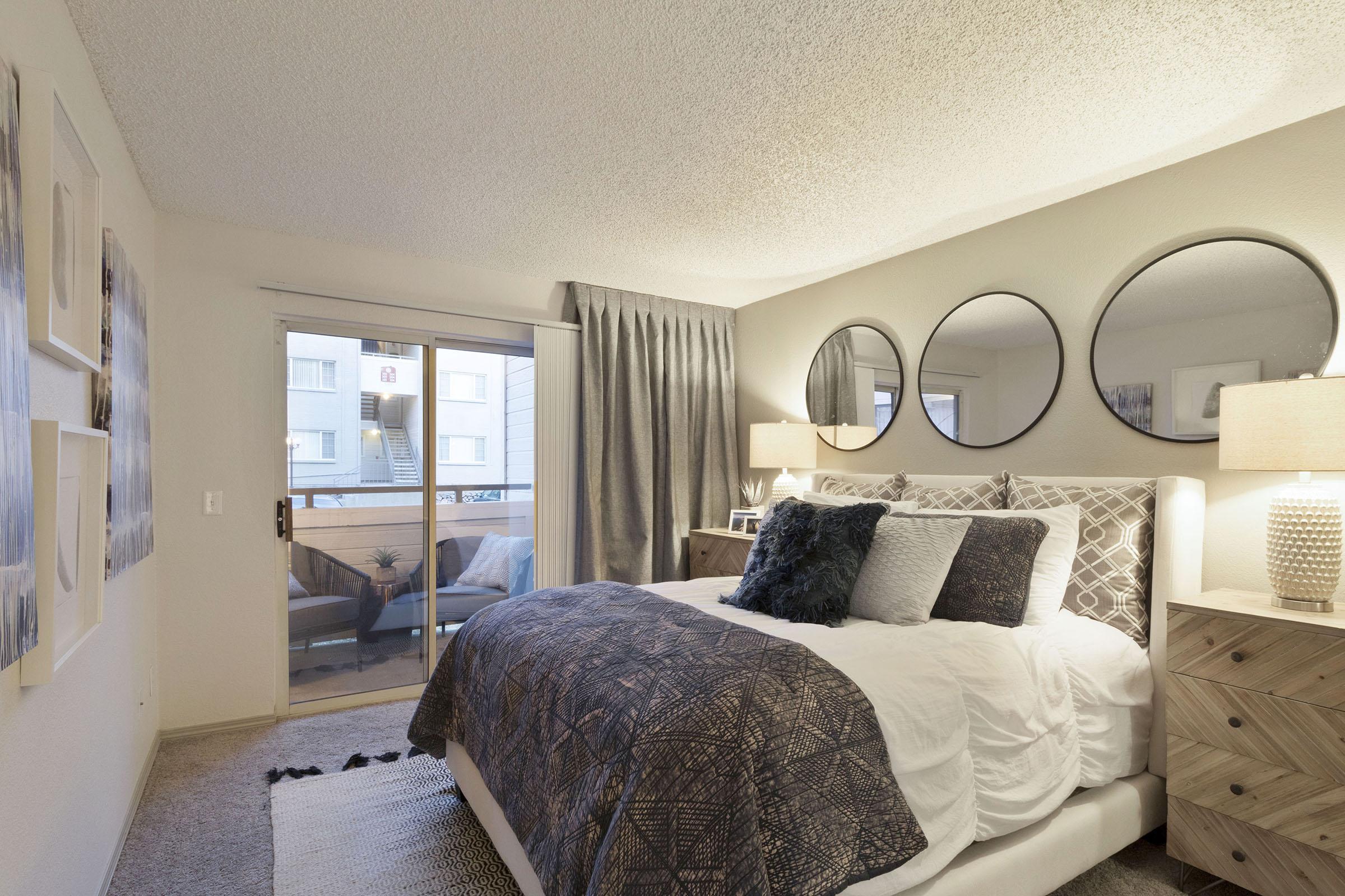 A cozy bedroom featuring a large bed with decorative pillows and a textured blanket. The walls are adorned with modern art, and there are three round mirrors above the bed. Two lamps provide soft lighting, and a balcony is visible through sliding glass doors, with sheer curtains enhancing the ambiance.