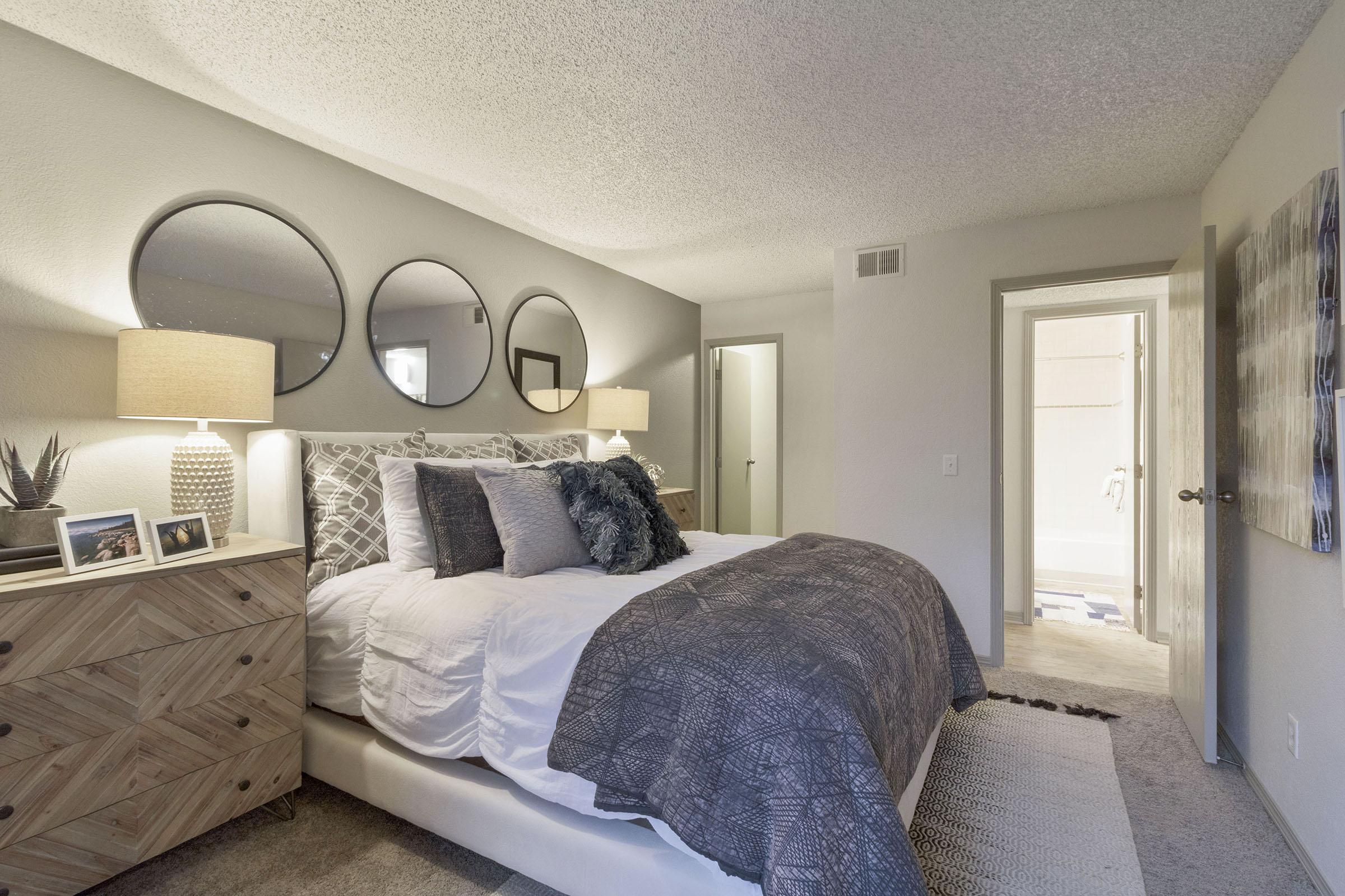 A stylish bedroom featuring a cozy bed with decorative pillows and a quilt, two round mirrors above the bed, and modern bedside lamps. A textured nightstand adds warmth, while a door leads to an adjacent bathroom. The walls are a soft gray, creating a calm atmosphere.