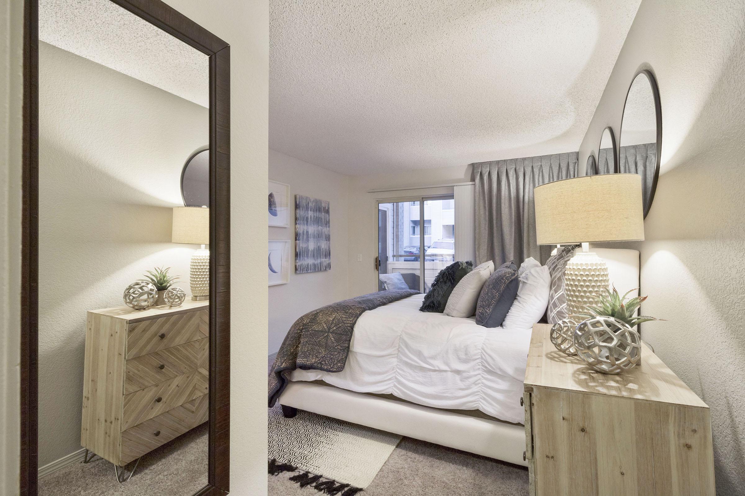 A cozy bedroom featuring a comfortable bed with white bedding and decorative pillows. A bedside table with a stylish lamp and decorative accents sits next to the bed. A large mirror reflects the room, and light-colored curtains frame a window. Artwork adorns the walls, adding a touch of personality to the space.