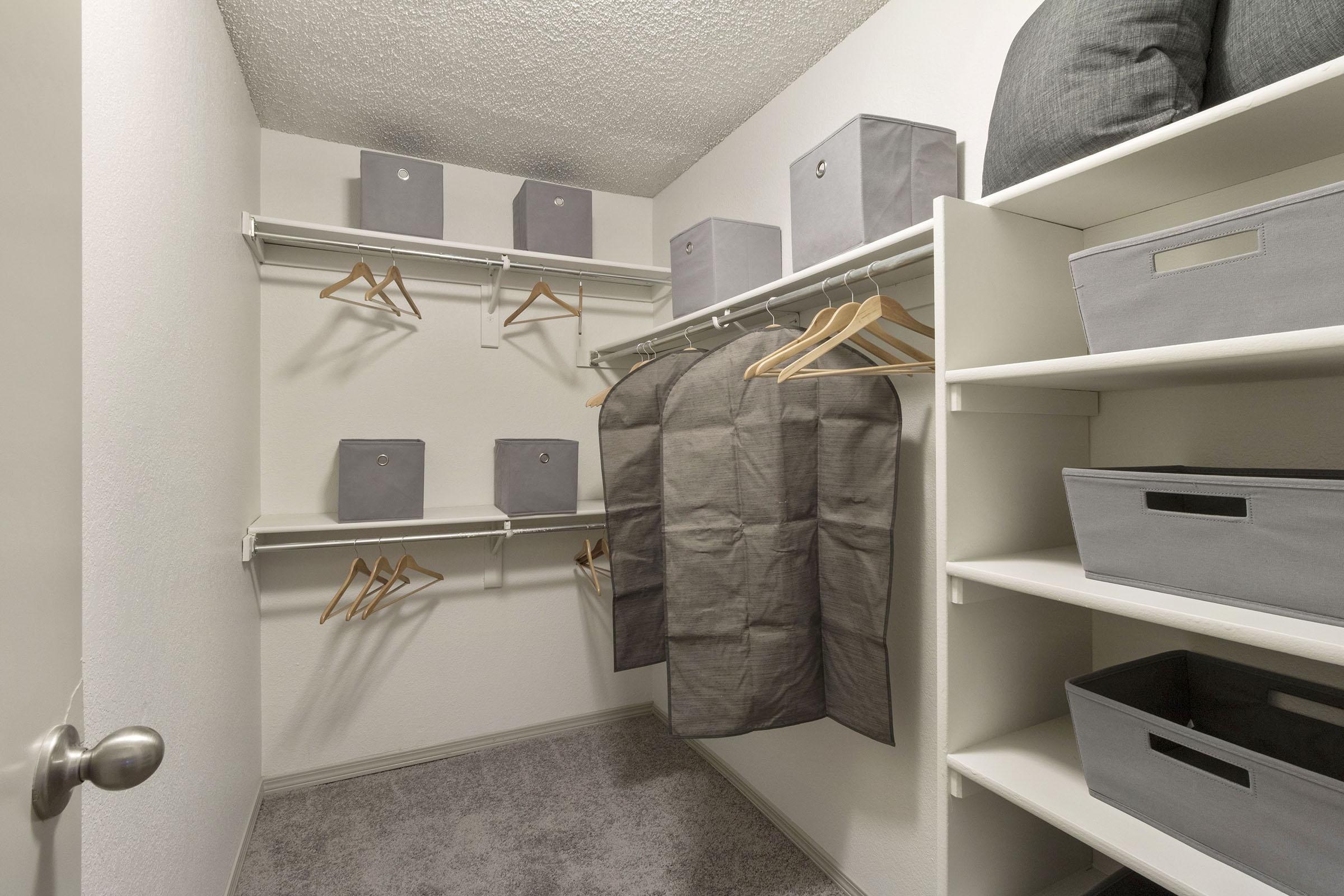 A neatly organized closet featuring light gray storage boxes, hanging clothes, wooden hangers, and shelves with additional gray storage bins. The walls are painted white, and the flooring is carpeted in a light shade. The overall appearance is clean and minimalistic.