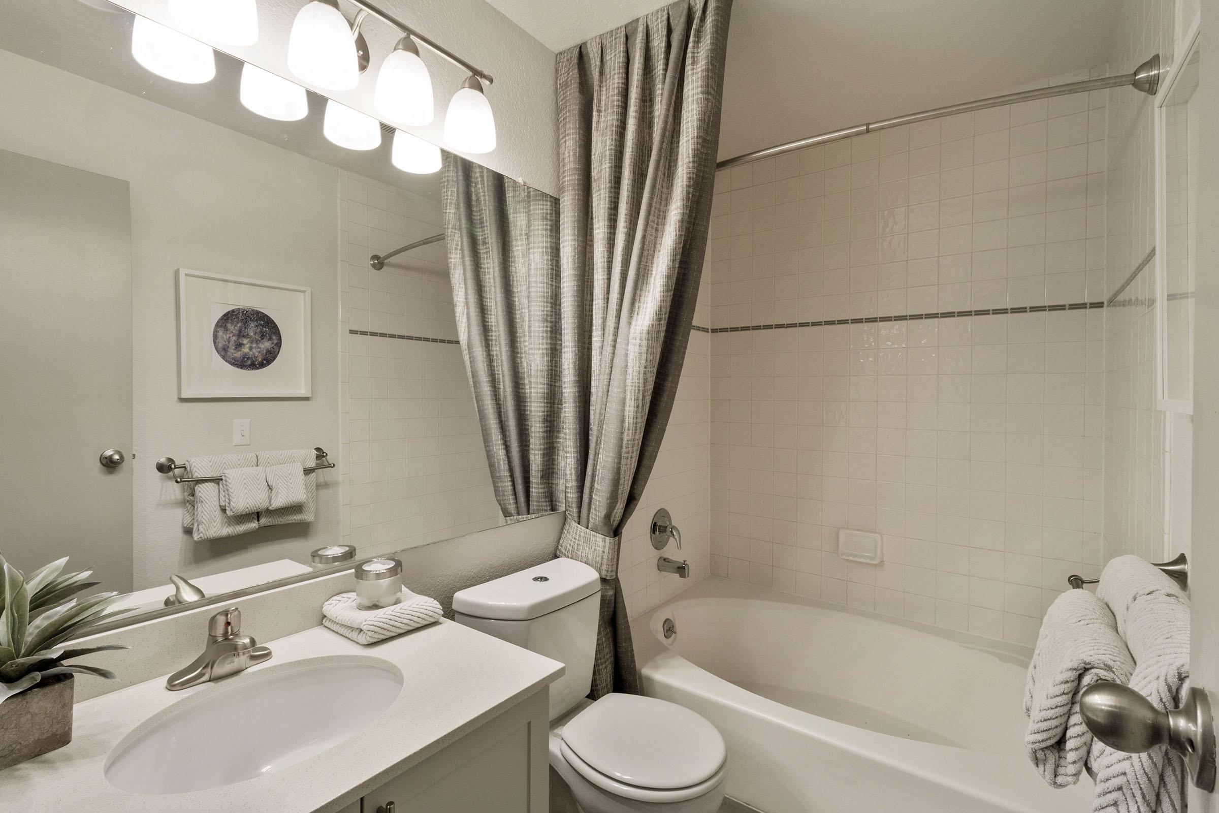 A modern bathroom featuring a white tub with a shower curtain, a lighted mirror above a sink with a vanity, and neatly arranged towels. A small plant decorates the counter, and the overall color scheme is neutral with soft lighting, creating a clean and inviting atmosphere.