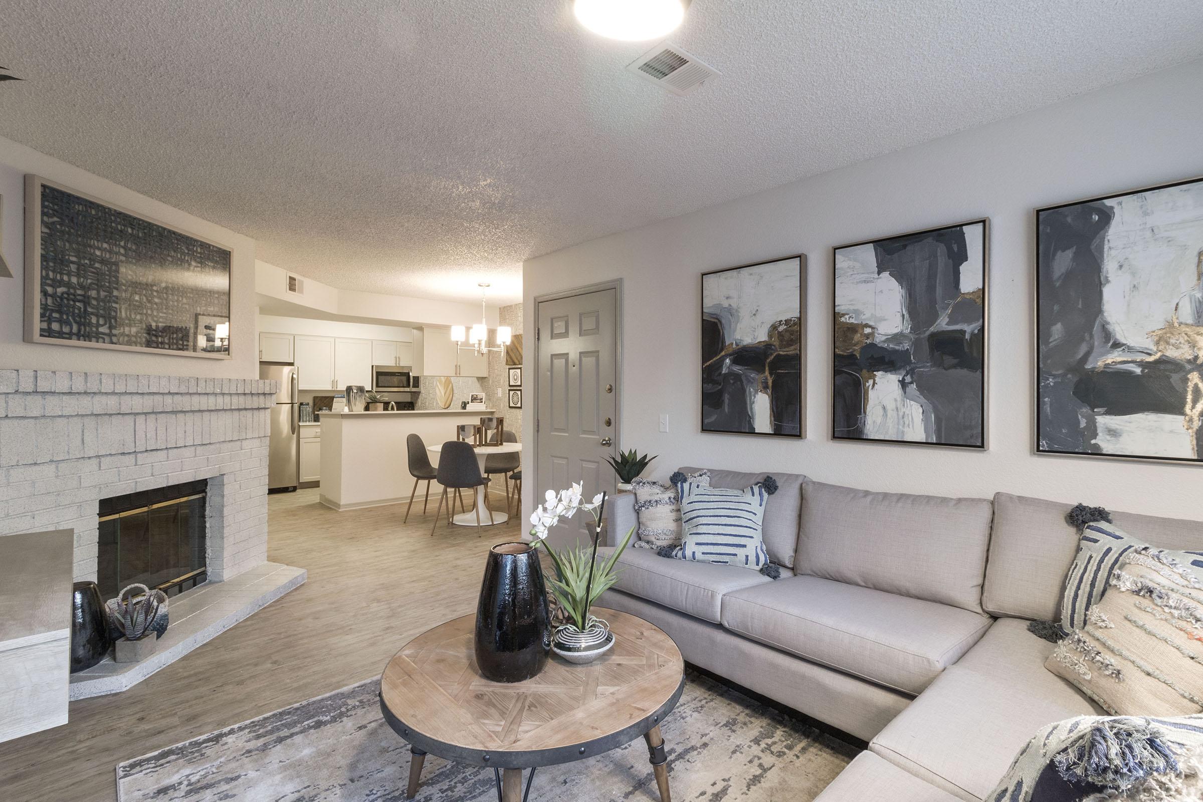 A modern living room featuring a light gray sectional sofa, a round coffee table, and a fireplace. Abstract artwork adorns the walls, and there’s a vase with white flowers on the table. In the background, a glimpse of a kitchen area can be seen, creating a cozy and inviting atmosphere.