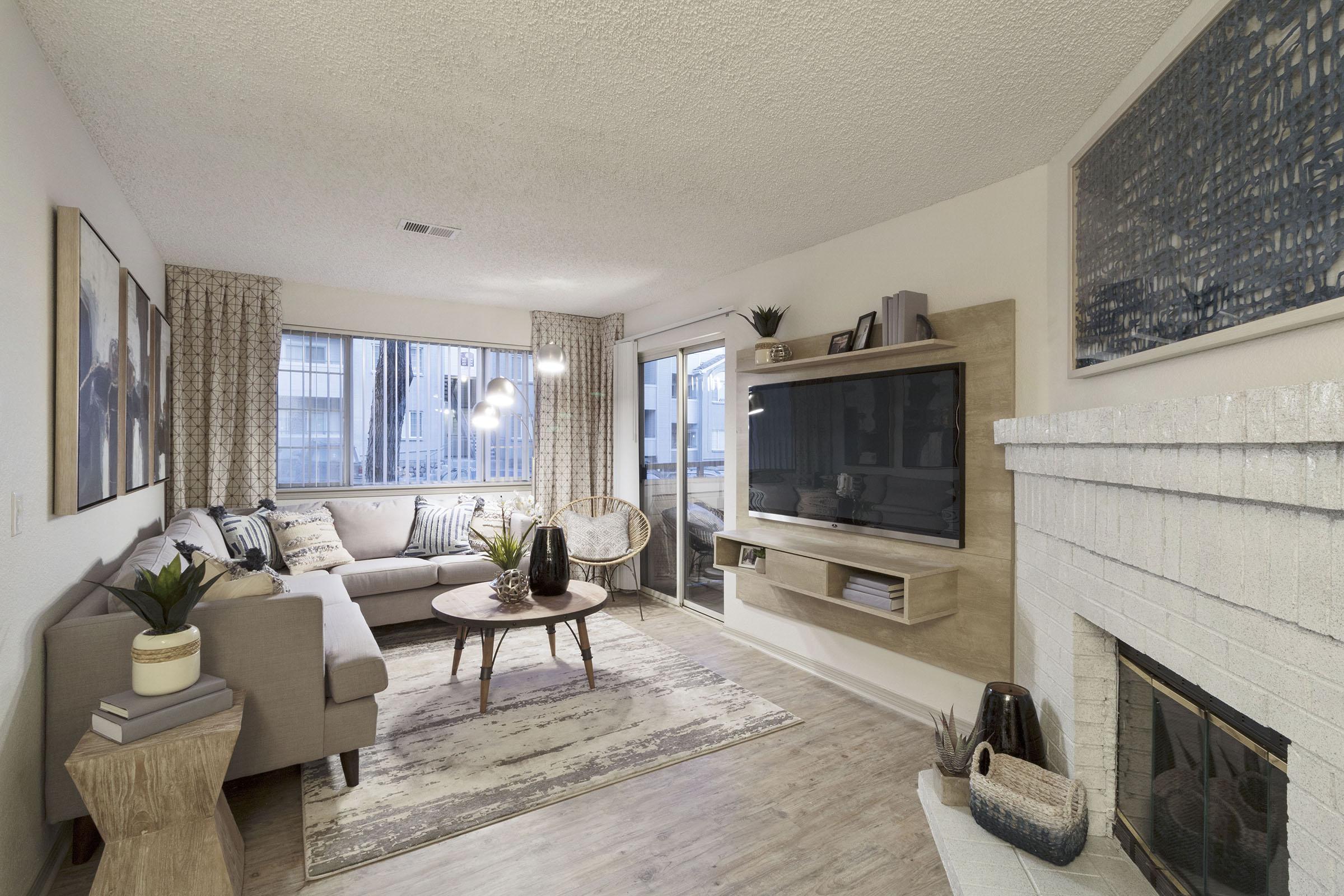 A modern living room with a light color scheme. It features a grey sofa, a round coffee table, and a decorative rug. There's a wall-mounted TV, a stone fireplace, and large windows with sheer curtains. Decorative plants and art pieces enhance the cozy ambiance.