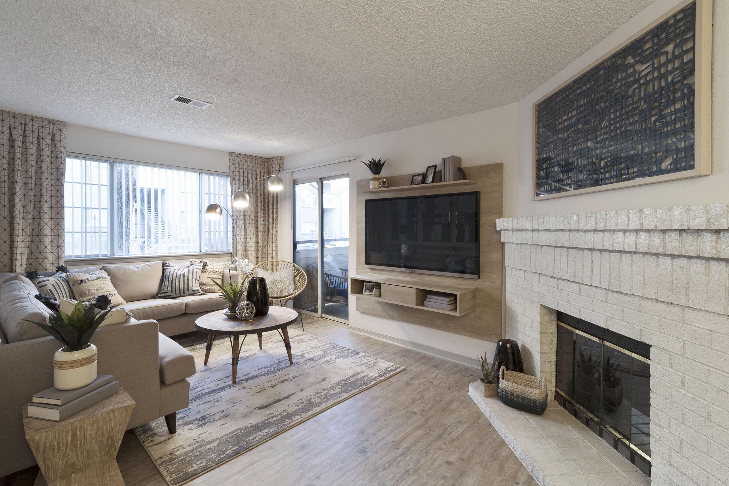 A cozy living room featuring a light-colored sofa, round coffee table, and textured area rug. Large windows allow natural light, and a fireplace with a stone facade adds warmth. A mounted TV sits on a wooden wall unit, and decorative plants enhance the inviting atmosphere.