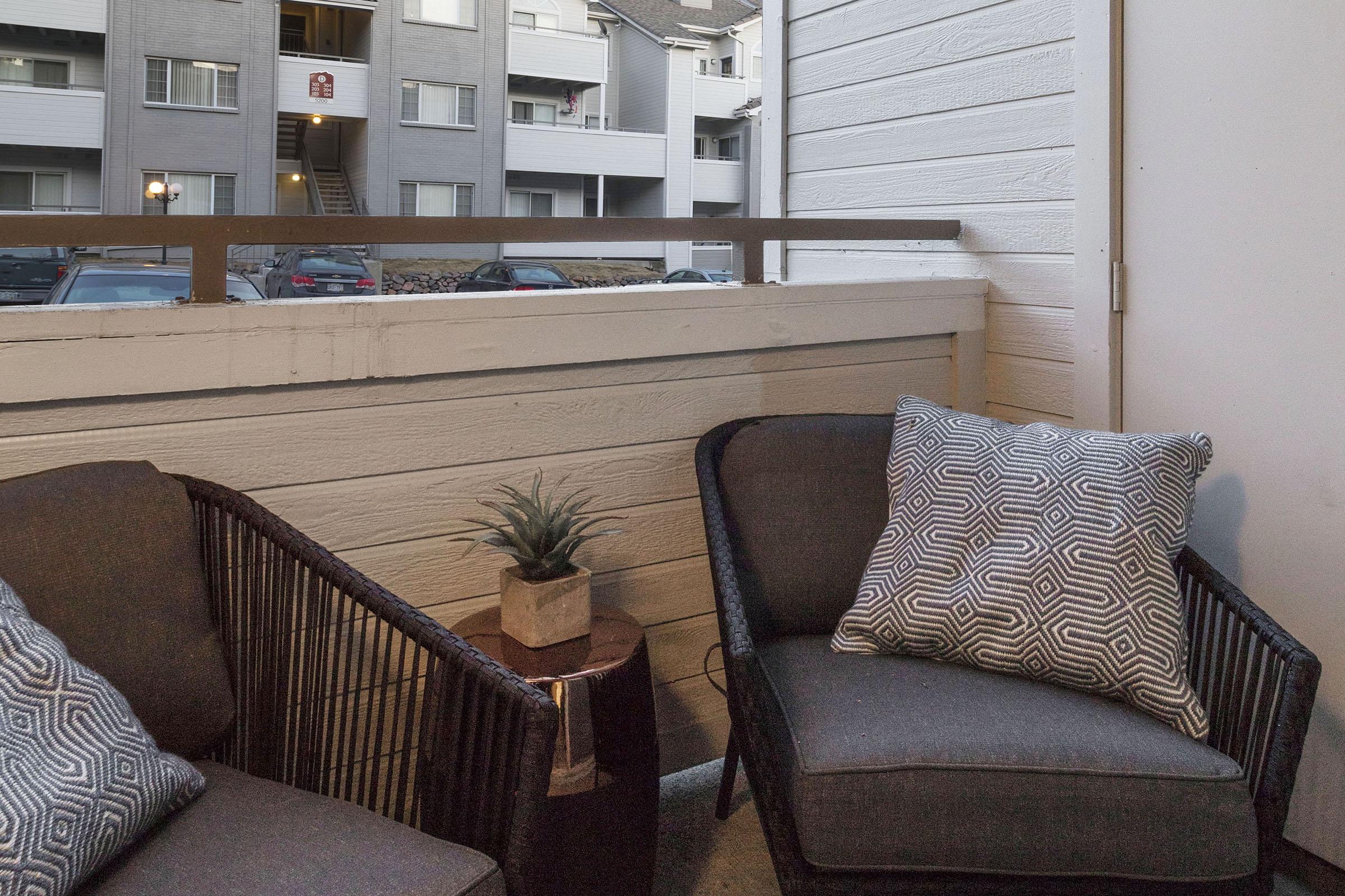 A cozy balcony space featuring two woven chairs with decorative cushions, a small side table with a plant on it, and a view of an apartment building across the way. The setting suggests a welcoming outdoor retreat.