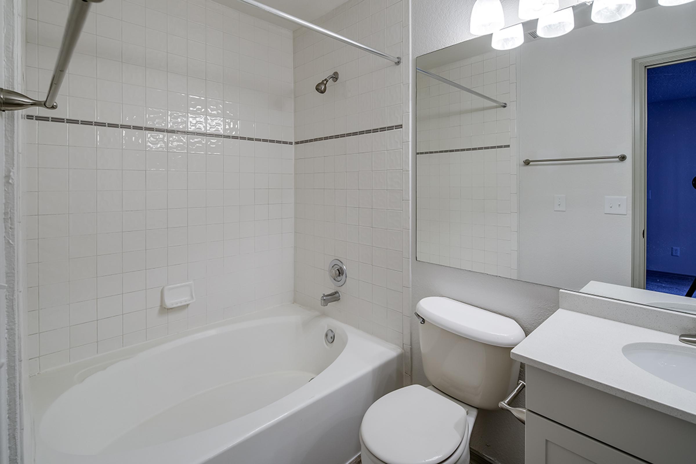 A clean and modern bathroom featuring a white tiled shower and bathtub, a toilet, and a sink with a countertop. The space is well-lit with overhead lighting and a large mirror on the wall, creating a bright and airy atmosphere. The overall design is minimalist and functional.