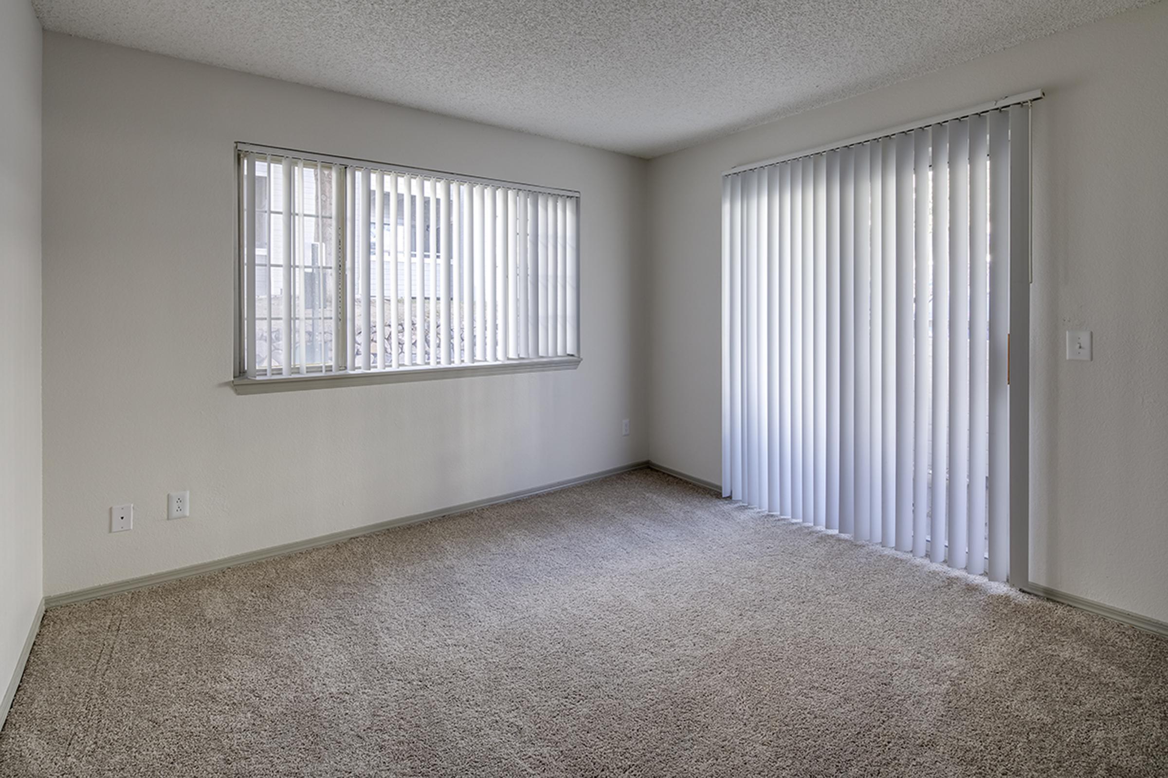 A minimalist interior of a room featuring beige carpeted flooring and pale walls. There are two windows: one with slatted blinds and the other with vertical blinds. The space is empty, creating a bright and airy atmosphere with natural light streaming in.