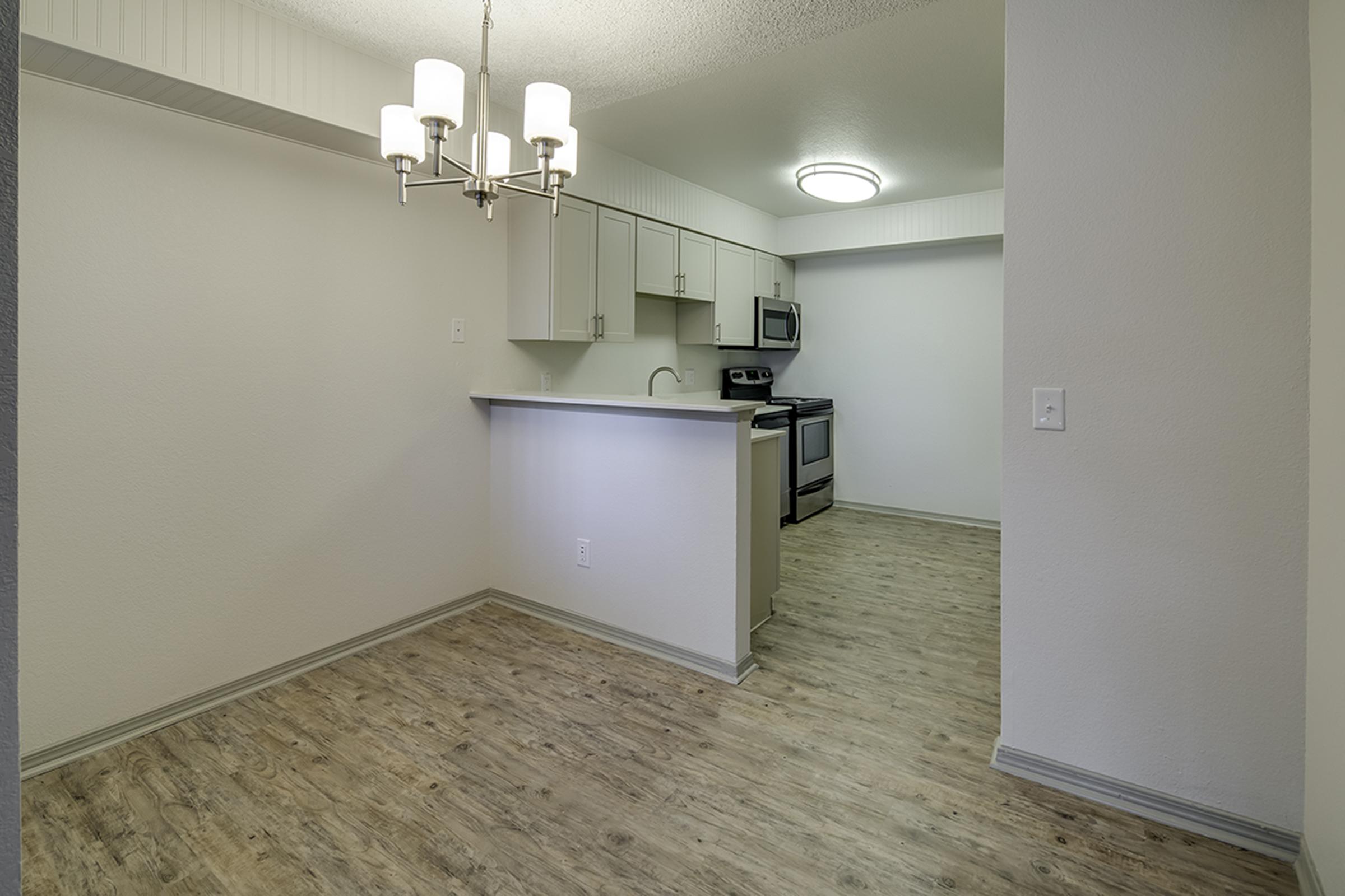 A modern kitchen and dining area featuring light wood flooring, white cabinetry, and stainless steel appliances. A chandelier hangs above an island counter, creating a bright and open space. The walls are painted in neutral tones, enhancing the contemporary look of the room.