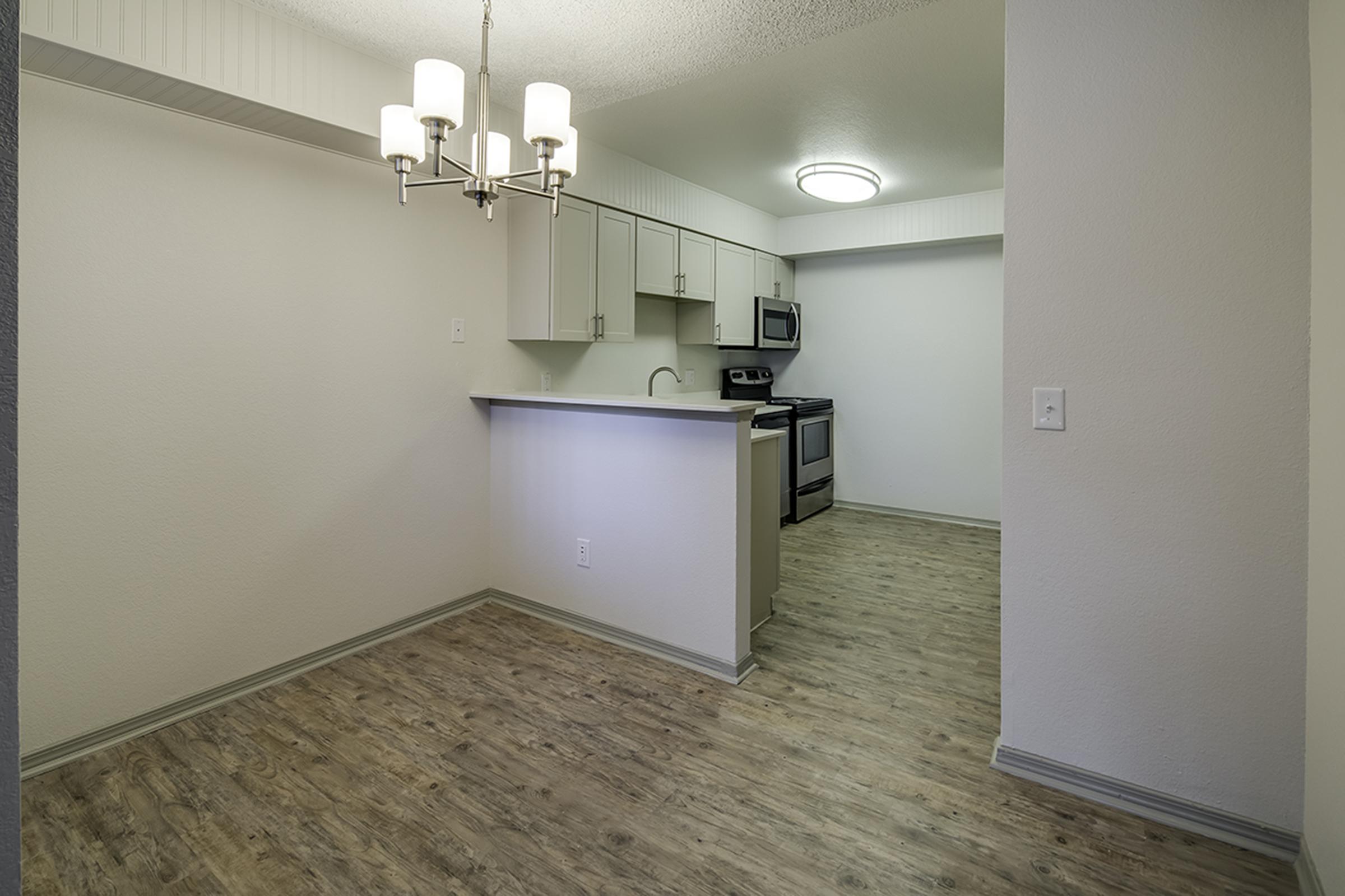 A modern kitchen and dining area featuring light wood flooring, a grey kitchen island with a sink, white cabinets, stainless steel appliances, and a chandelier hanging from the ceiling. The walls are painted in neutral tones, creating a bright and airy atmosphere.