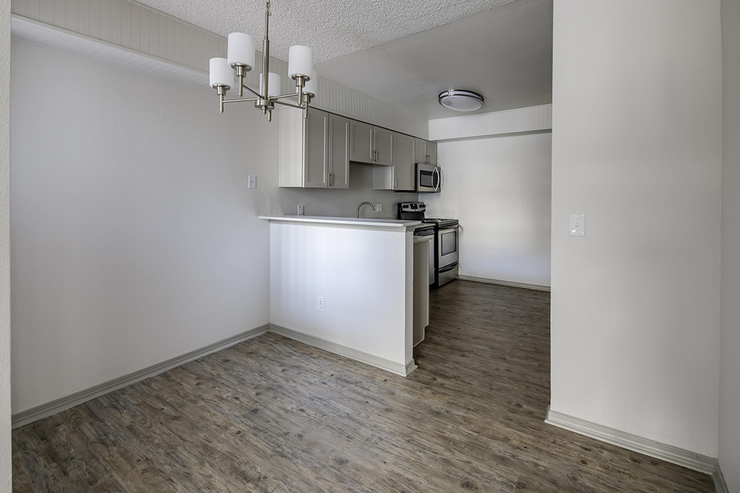 A bright, modern kitchen and dining area with light wood flooring. The kitchen features white cabinets, stainless steel appliances, and a small island with a sink. Overhead, a chandelier and a ceiling light provide illumination. The room has an open layout with white walls and an airy feel.