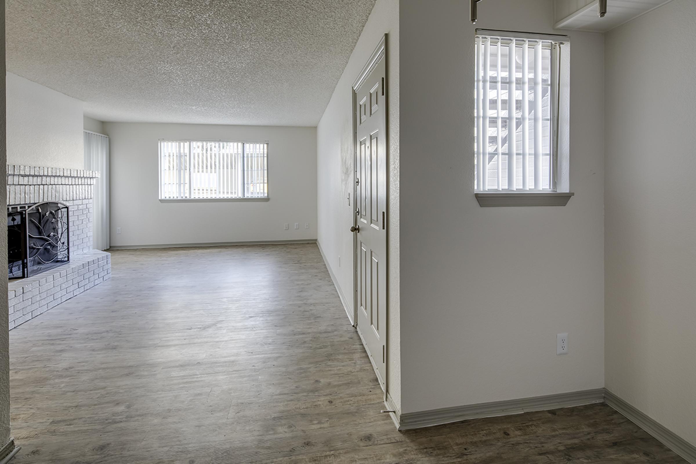 A spacious, well-lit interior of a modern apartment featuring a living area with light-colored walls and a textured ceiling. It includes large windows with blinds, a doorway with a door, and wood-like flooring. A fireplace is visible on the left side, enhancing the cozy atmosphere.