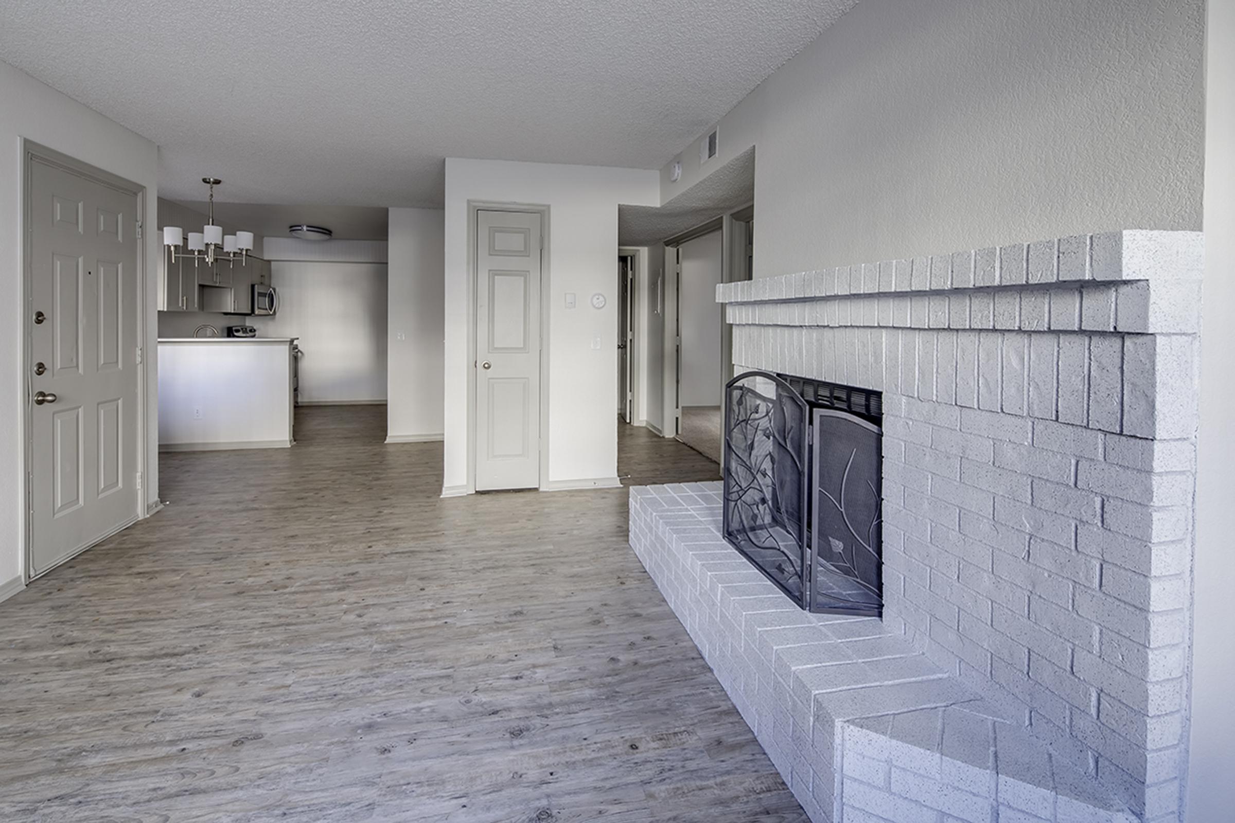 A spacious, modern living room featuring light gray walls and light wood flooring. A brick fireplace is on the right side, while the kitchen and dining area are visible in the background, equipped with overhead lighting and a neutral color palette. Doorways lead to other rooms in the apartment.